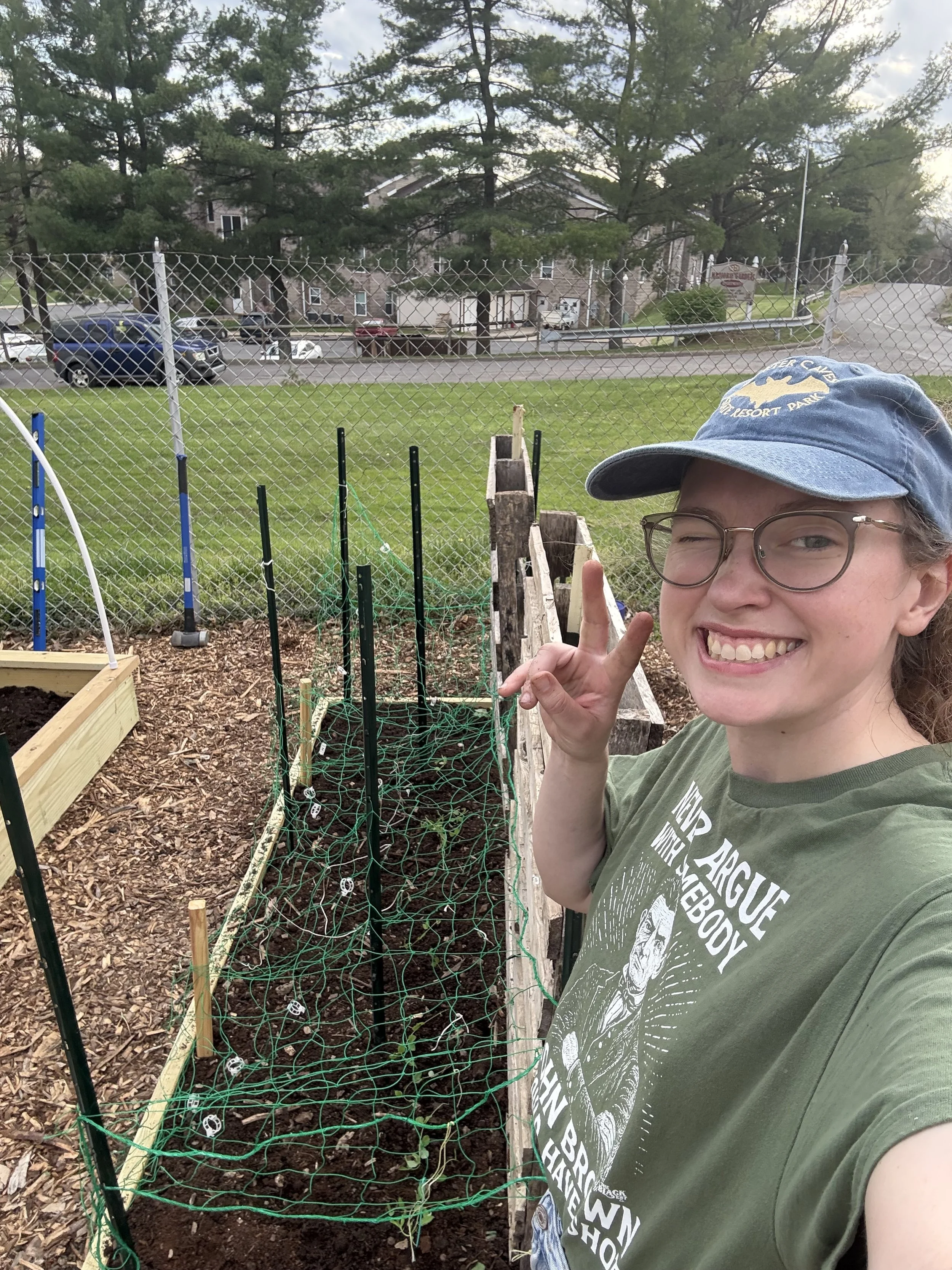 Paige Reiring gives a peace sign in front of bed of bean plants at a community garden.