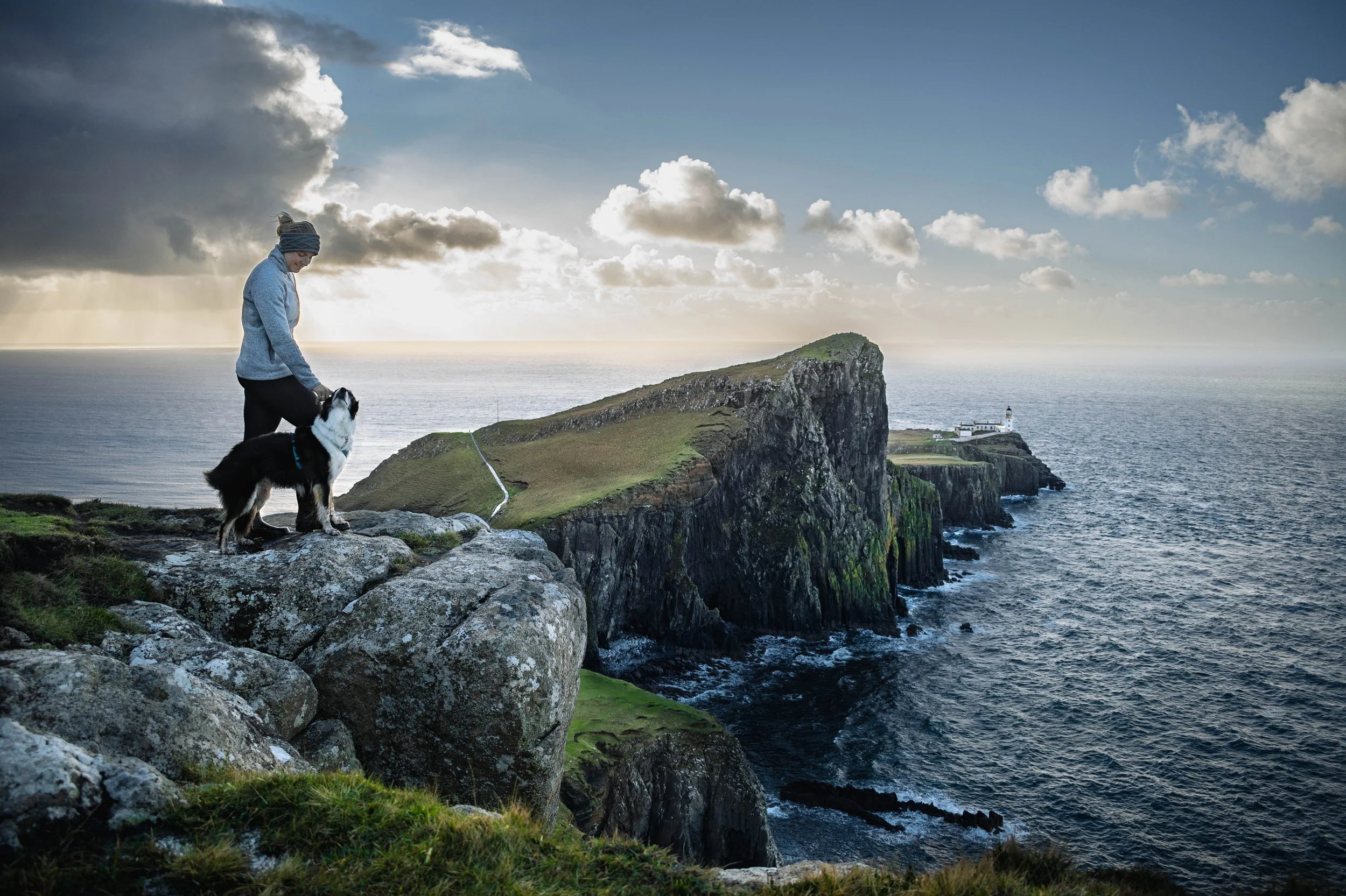 Eine Frau mit einem Hund steht auf felsigem Gelände an einer Klippe mit Blick auf das Meer. Im Hintergrund ist ein Leuchtturm auf einer kleinen Insel vor der schroffen Küste zu sehen, bei bewölktem Himmel.