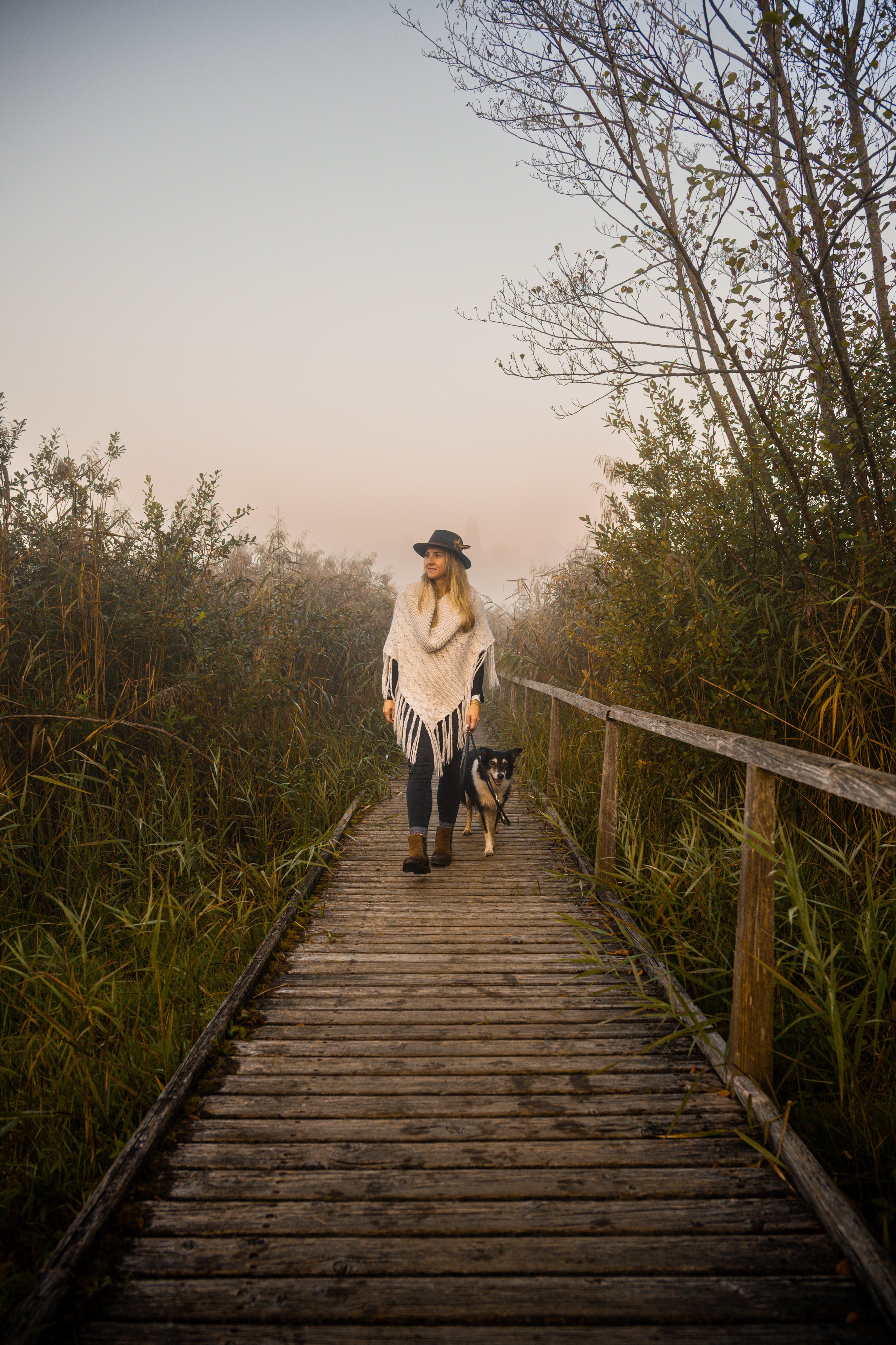 Frau mit Hut geht mit Hund auf einem holprigen Holzsteg durch eine neblige, bewachsene Landschaft.
