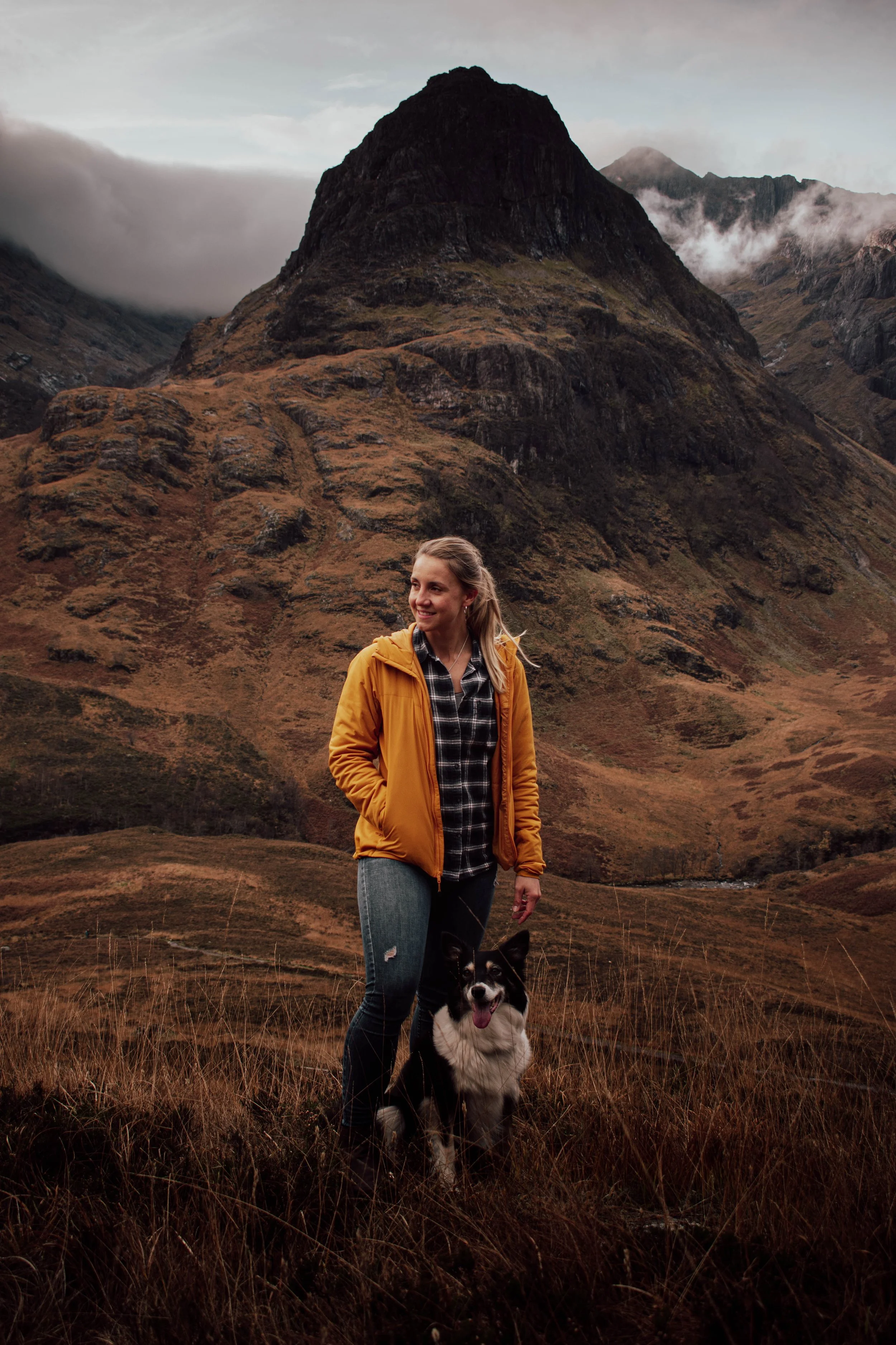 Eine junge Frau mit einem schwarzen und weißen Hund in einer ländlichen, bergigen Landschaft mit grauen Wolken am Himmel.