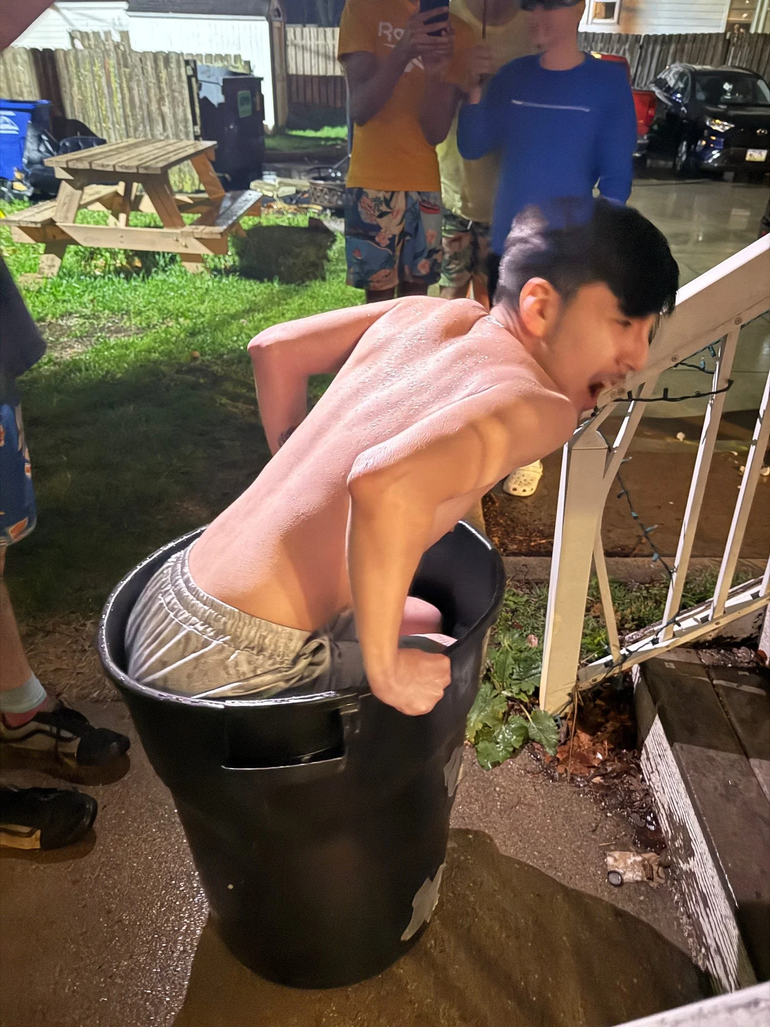 A shirtless man sitting in a large black trash can outdoors at night, laughing with his head tilted back and mouth open, with several people standing behind him.