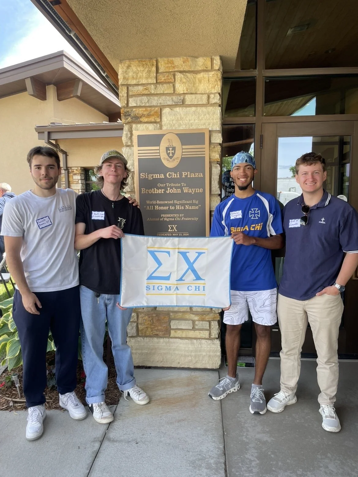 Four young men standing outside in front of a stone wall and windows, holding a Sigma Chi fraternity banner and a plaque commemorating Brother John Wayne, with some wearing Sigma Chi clothing and name tags.