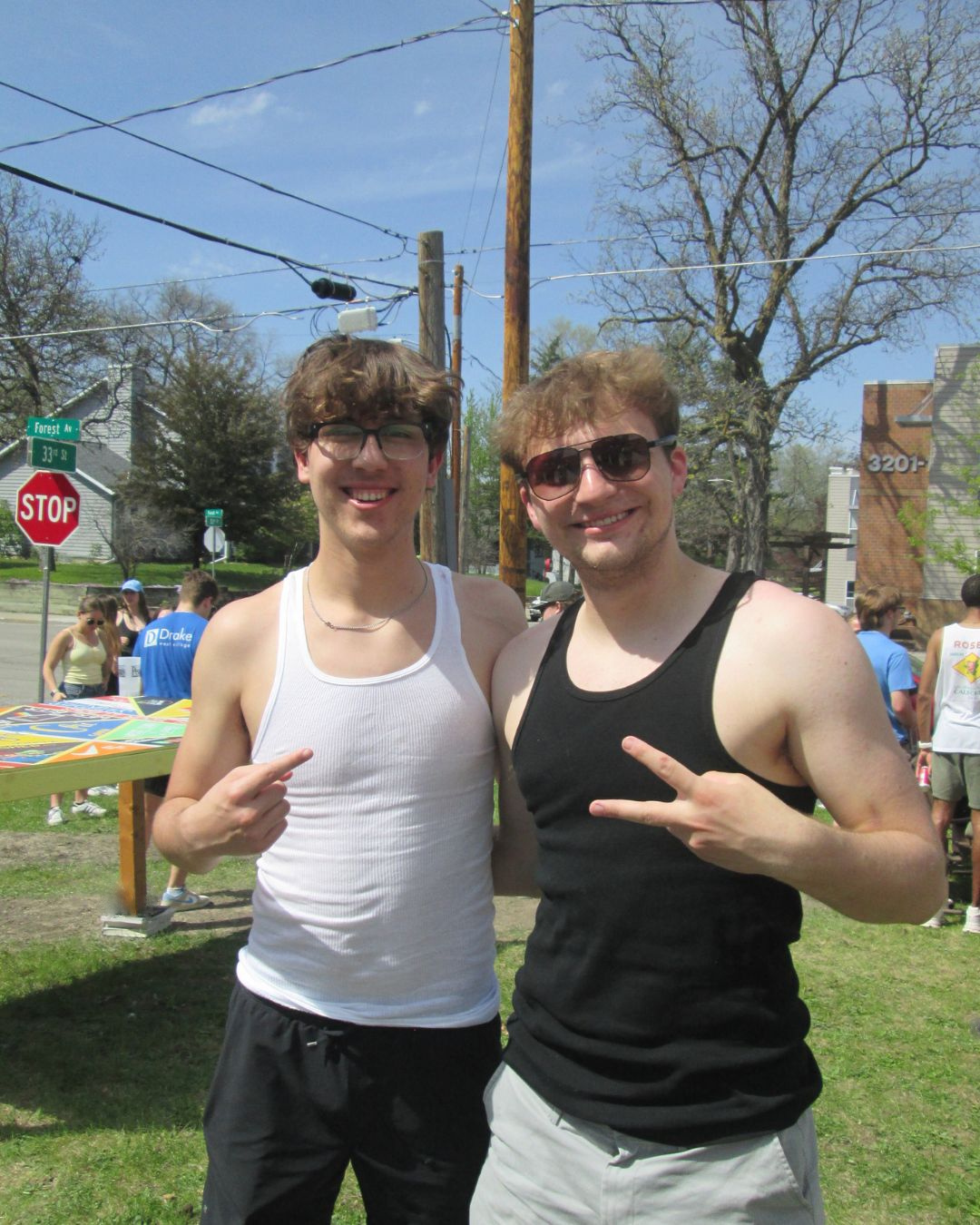 Two young men smiling outdoors, making peace signs with their hands during a sunny day at a street event or festival. One wears a white tank top and glasses, the other a black tank top and sunglasses.