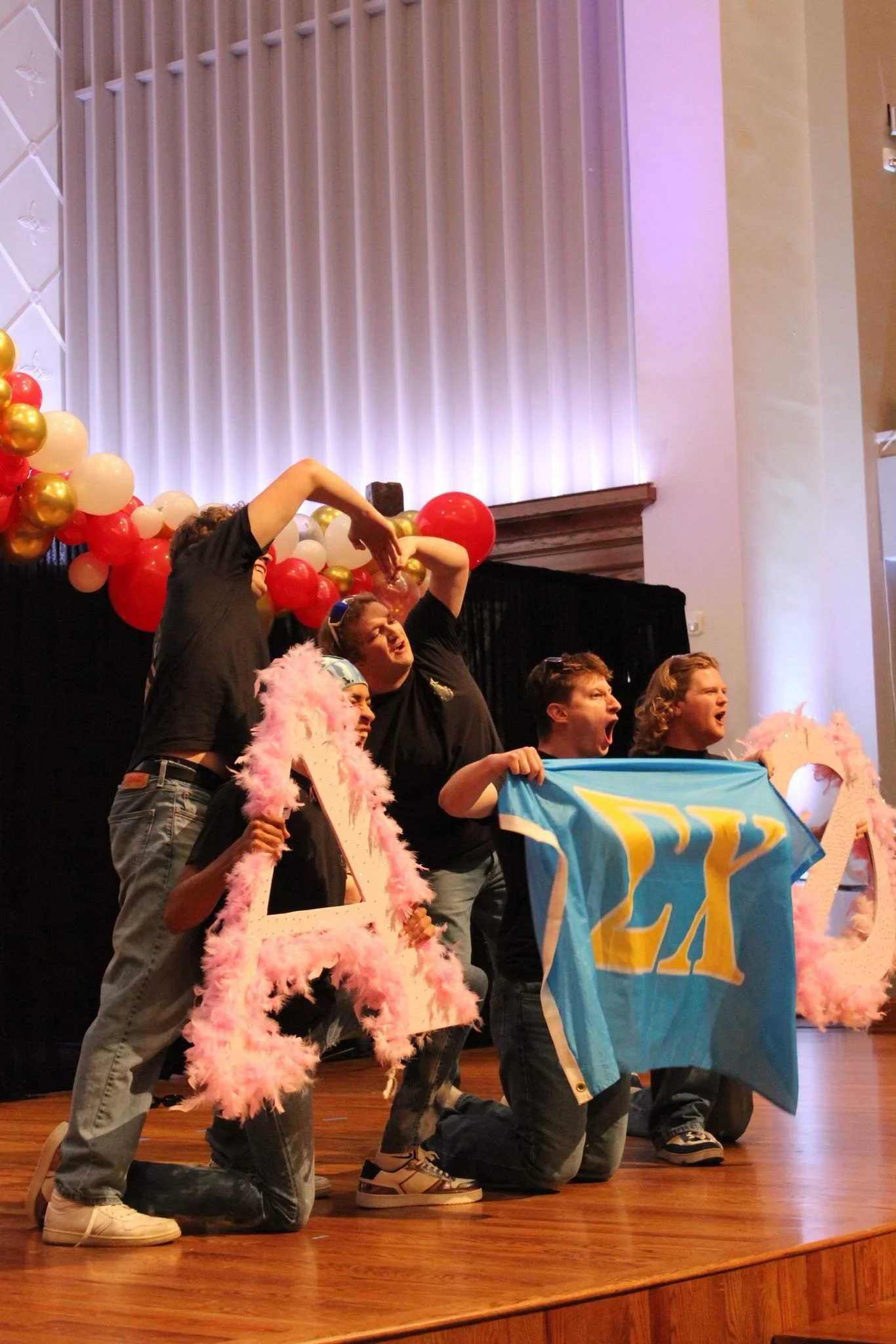 People on stage kneeling and holding flags, surrounded by balloons at an indoor event.