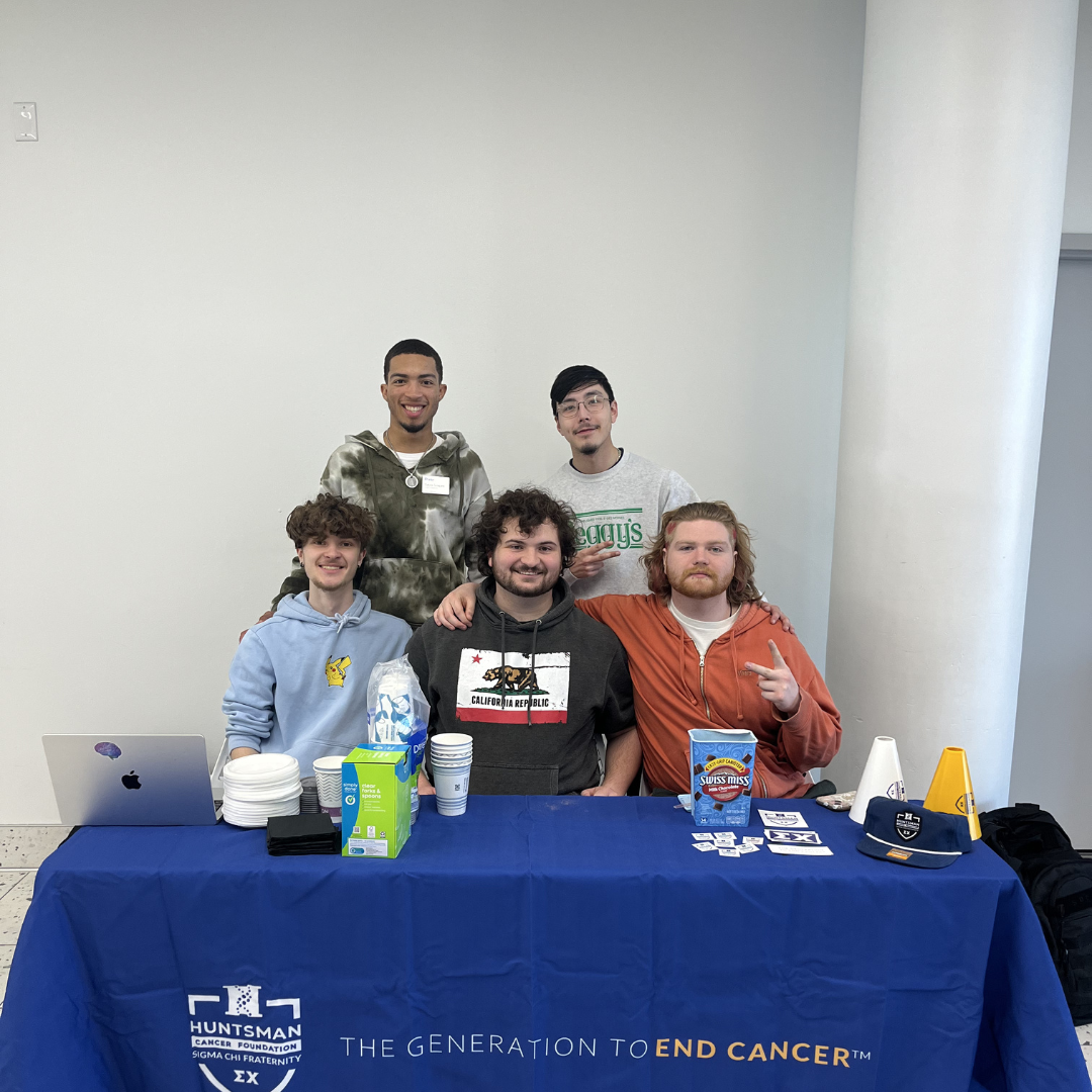 Group of five young men at a table for Huntsman Cancer Foundation, with a blue tablecloth displaying their logo and slogan, in a room with plain white walls.