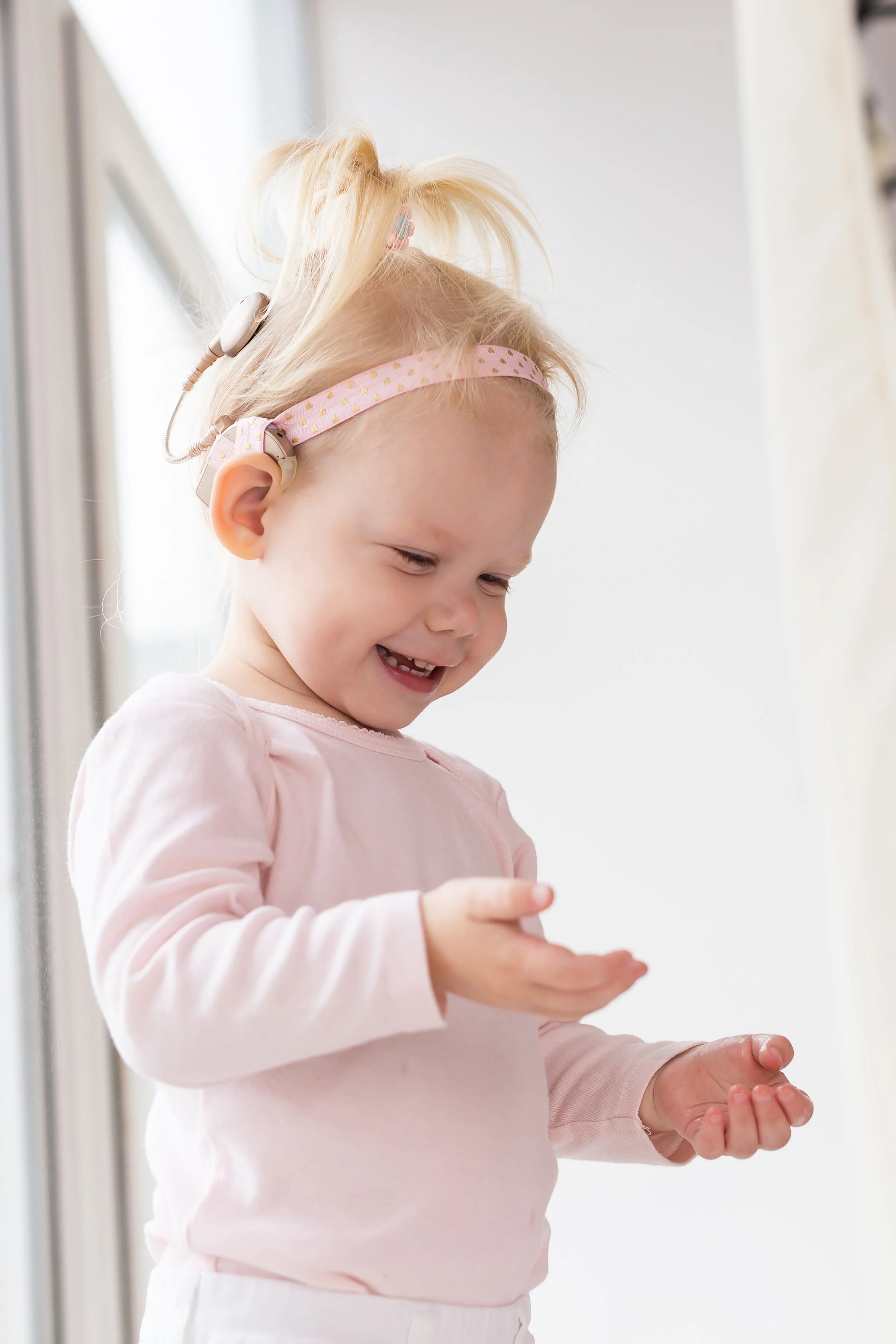 A young girl with a cochlear implant, wearing a pink headband and pink long sleeve top, is smiling and clapping her hands, during Listening & Spoken Language Therapy in Olathe, KS