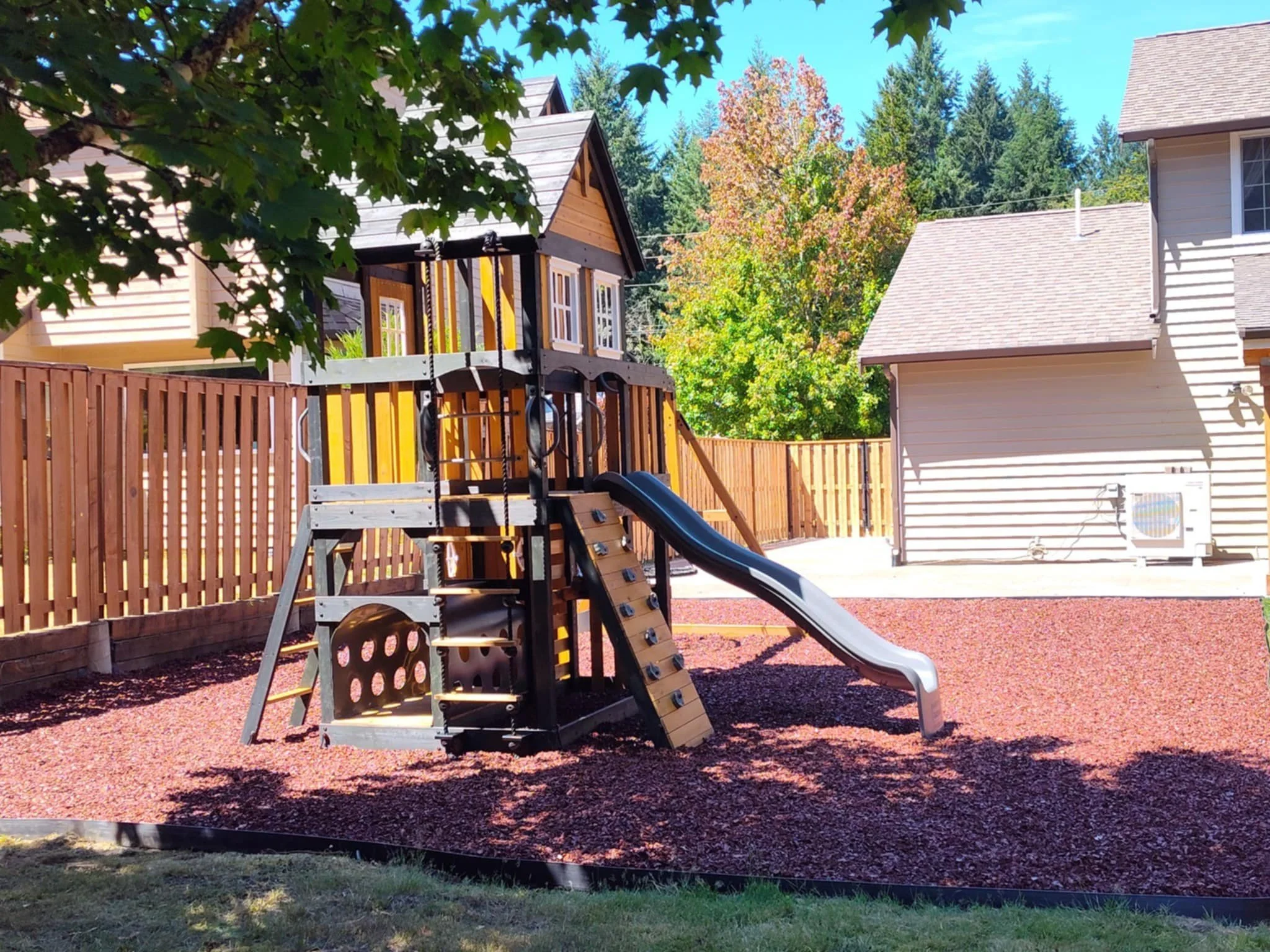 An outdoor two-level children's play structure with a roof and slide, surrounded by bark.