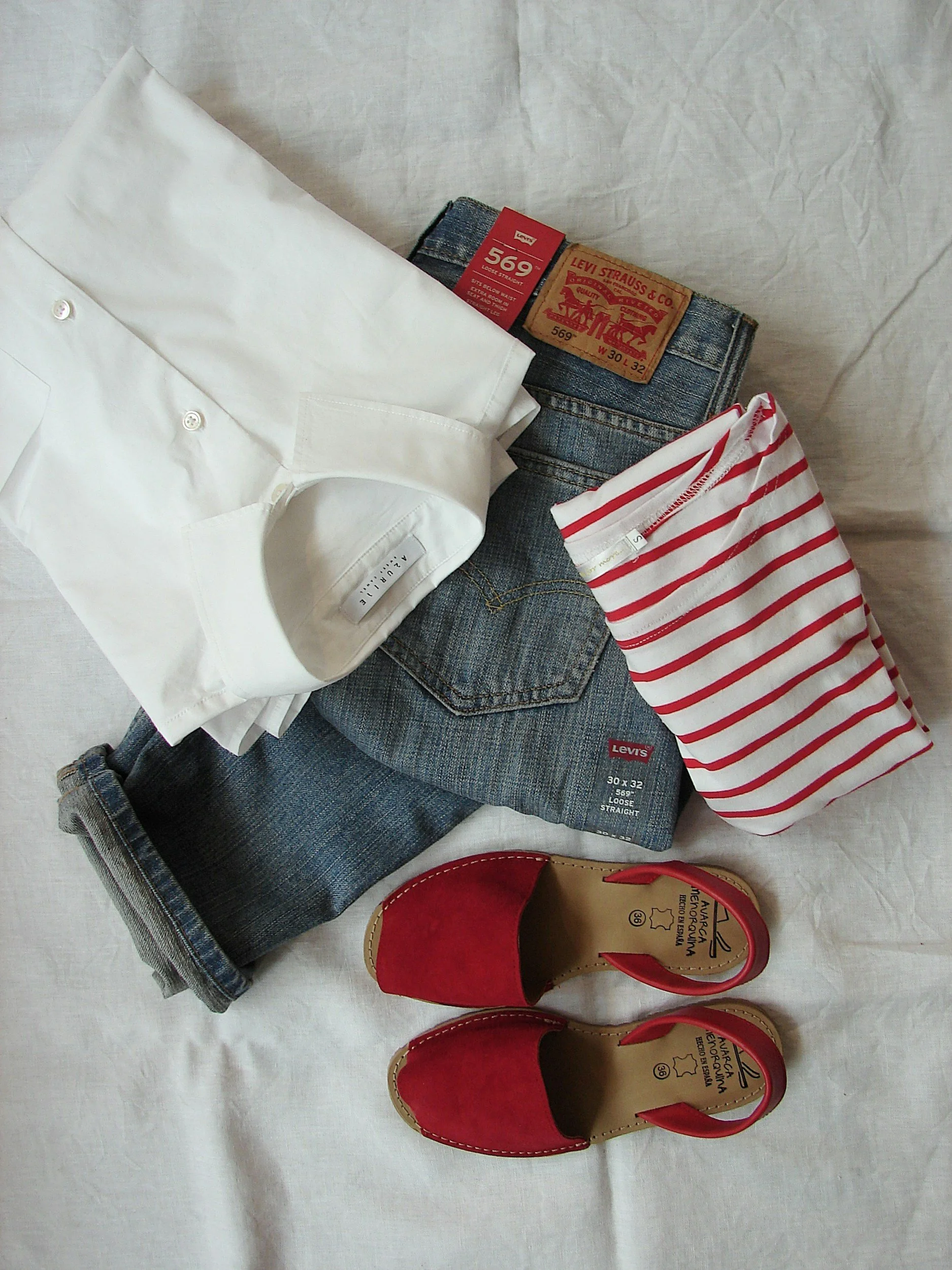 A white button-up shirt, a pair of blue Levi's jeans, a red and white striped shirt, and a pair of red pointed-toe shoes with beige soles and black text on the insoles are arranged on a light-colored surface.