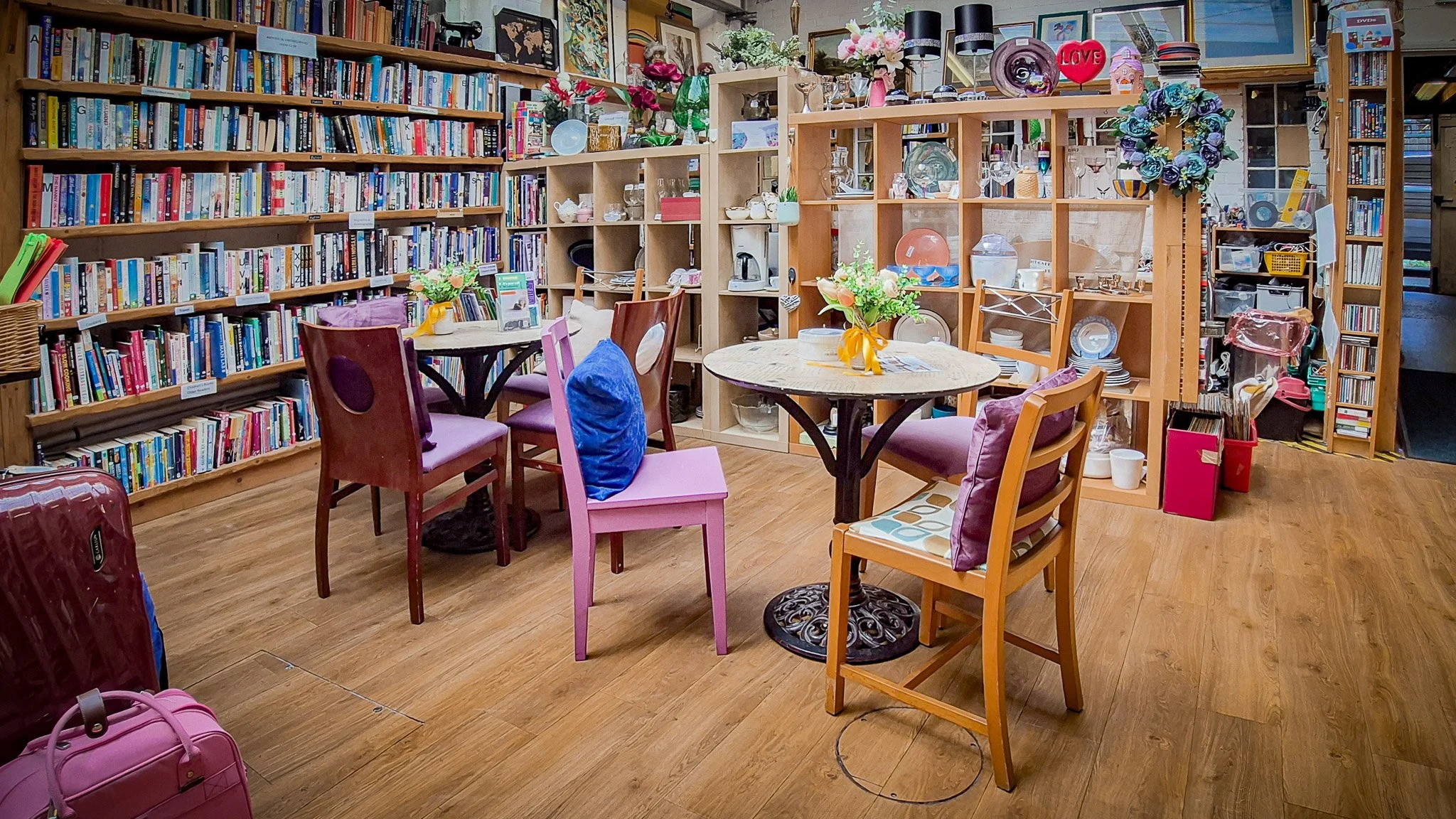 Interiort of a cozy bookstore or thrift shop with books on wooden shelves, round tables with flower arrangements, and a variety of chairs. Bright and colorful decor including a heart-shaped 'LOVE' sign and floral wreaths.