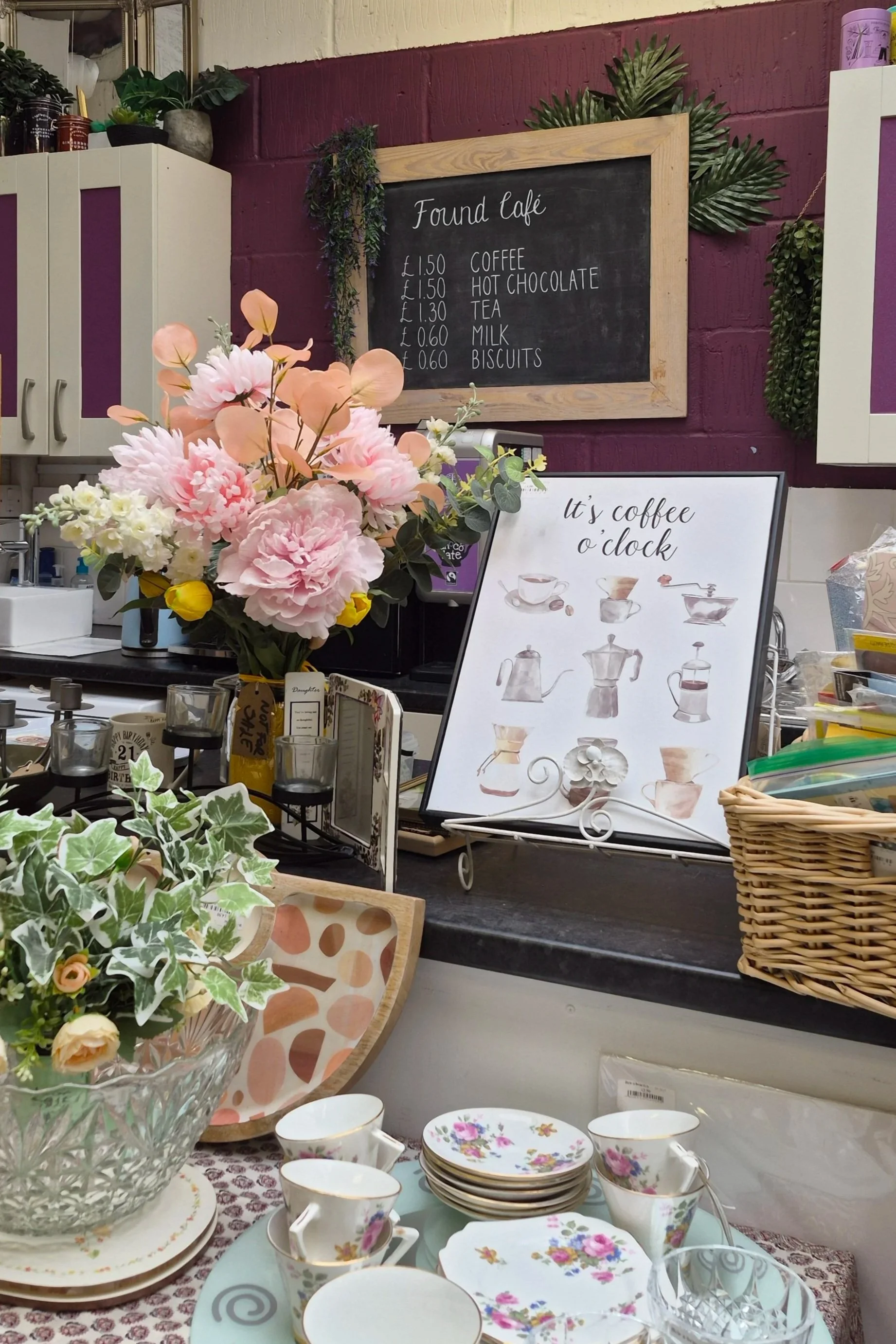 A display of floral china cups, saucers, and plates on a table, with a colorful bouquet of pink, peach, and white flowers in a glass vase. There is a basket on the right and decorative paper on the table. In the background, there is a description of coffee and tea on a framed chalkboard sign, and a smaller sign that says 'It's coffee o'clock'