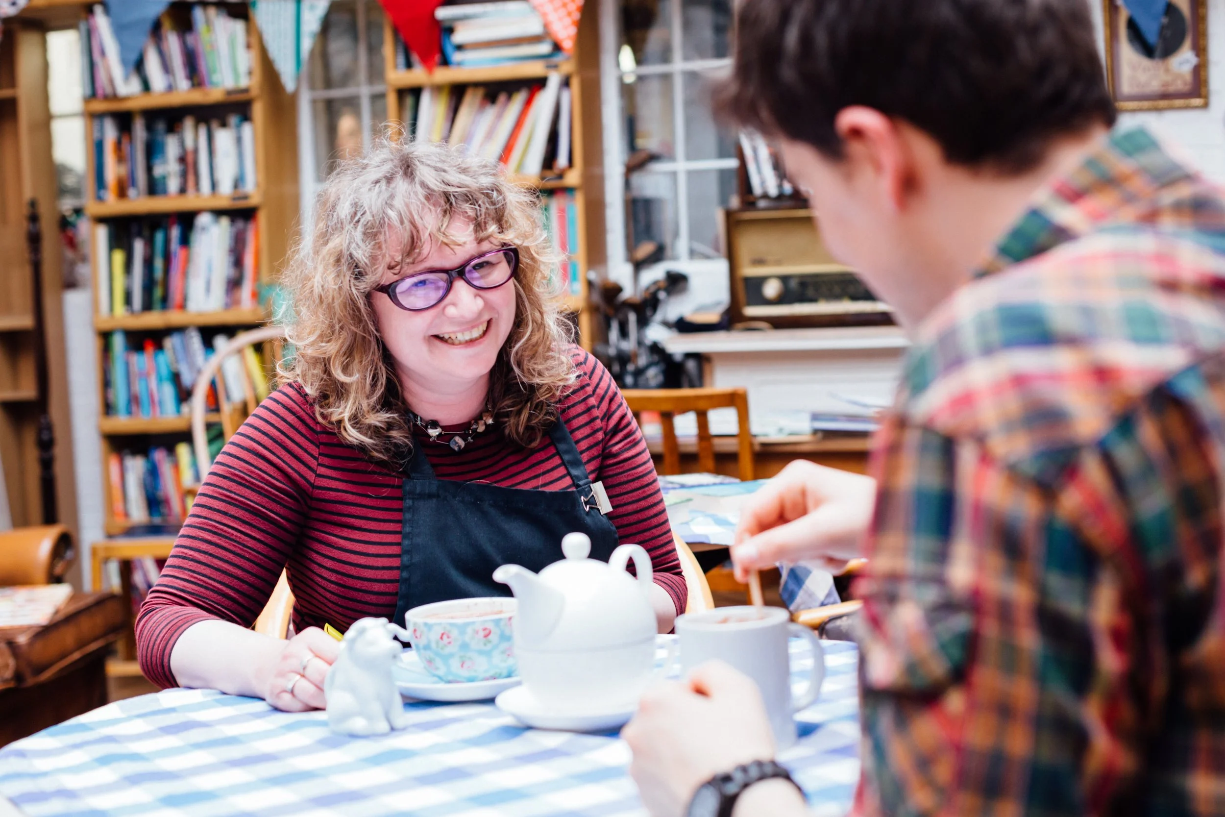 A woman with curly blonde hair and glasses smiling while sitting at a table during a tea party with a child. The table has a blue and white checkered tablecloth, teapot, cups, and decorative items. Bookshelves filled with books and a window are in the background.