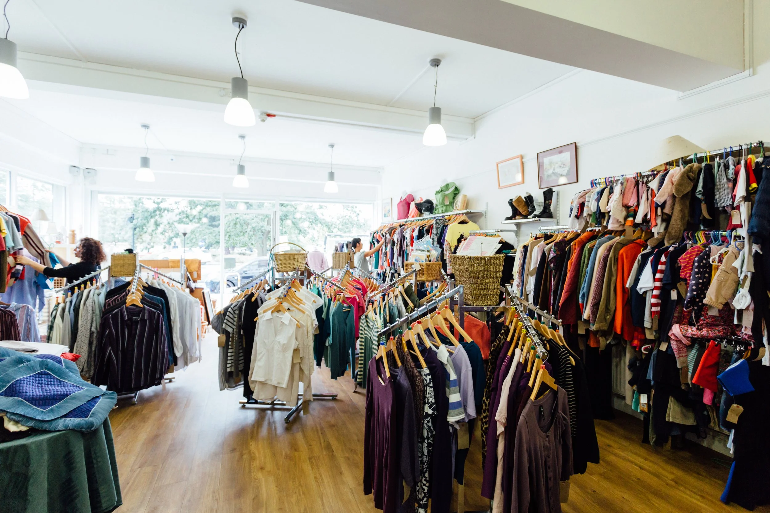 Interior view of a thrift store with racks of clothes including shirts, jackets, and sweaters, and a woman shopping near the windows with large windows letting in natural light.