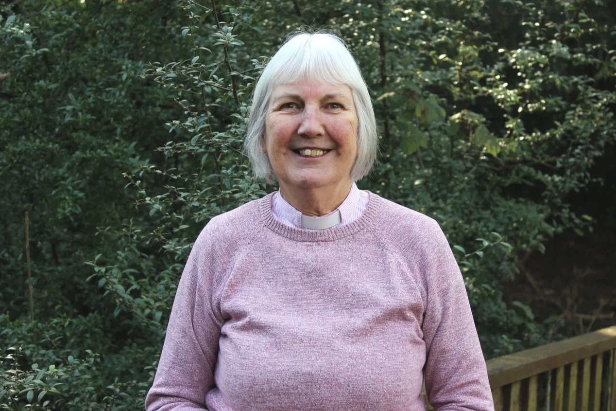 An older woman with white hair smiling outdoors in front of green bushes, wearing a pink sweater and a clerical collar.