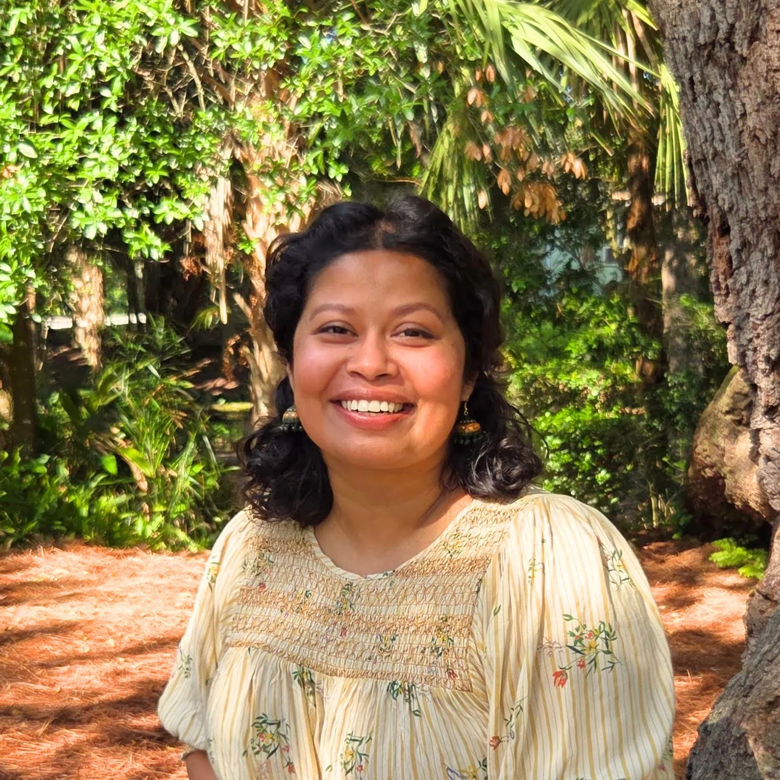 A woman with dark, curly hair smiling in a lush, green garden with a large tree trunk and dense foliage background.