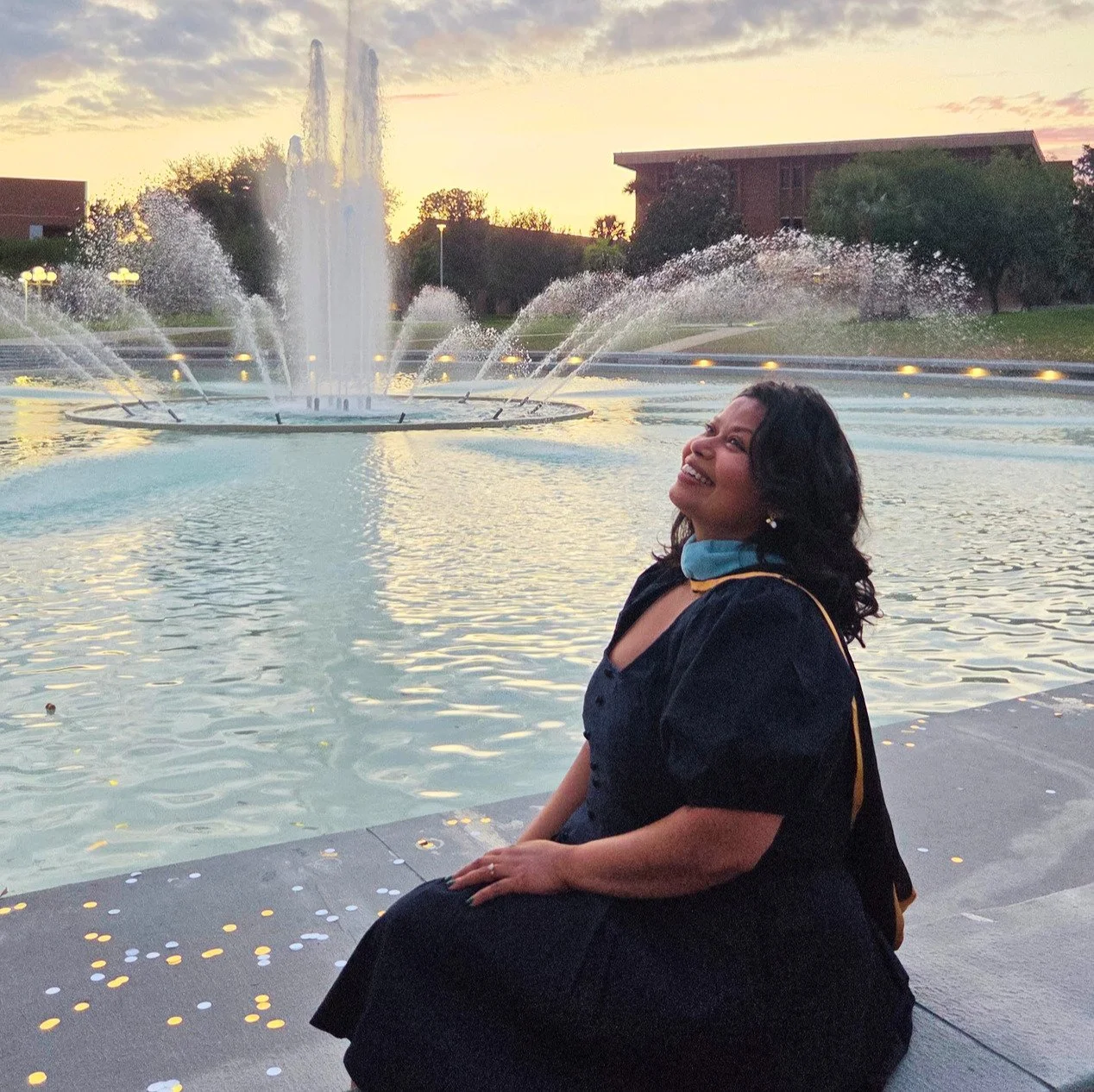 A woman in a graduation gown sits beside a fountain at sunset, smiling and looking upwards.