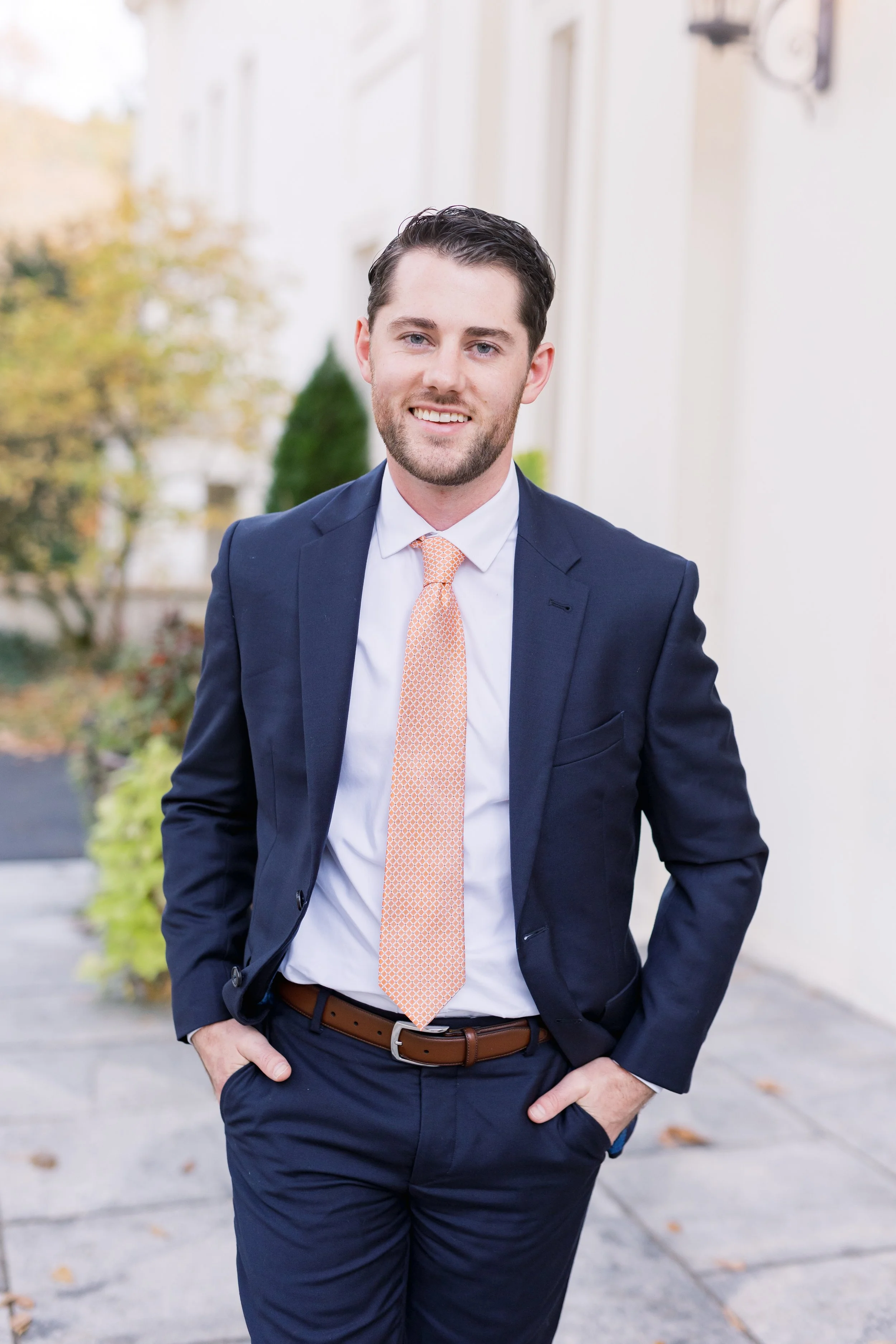 A young man in a navy blue suit with a light orange tie, standing outdoors with a slight smile, with trees and a building in the background.
