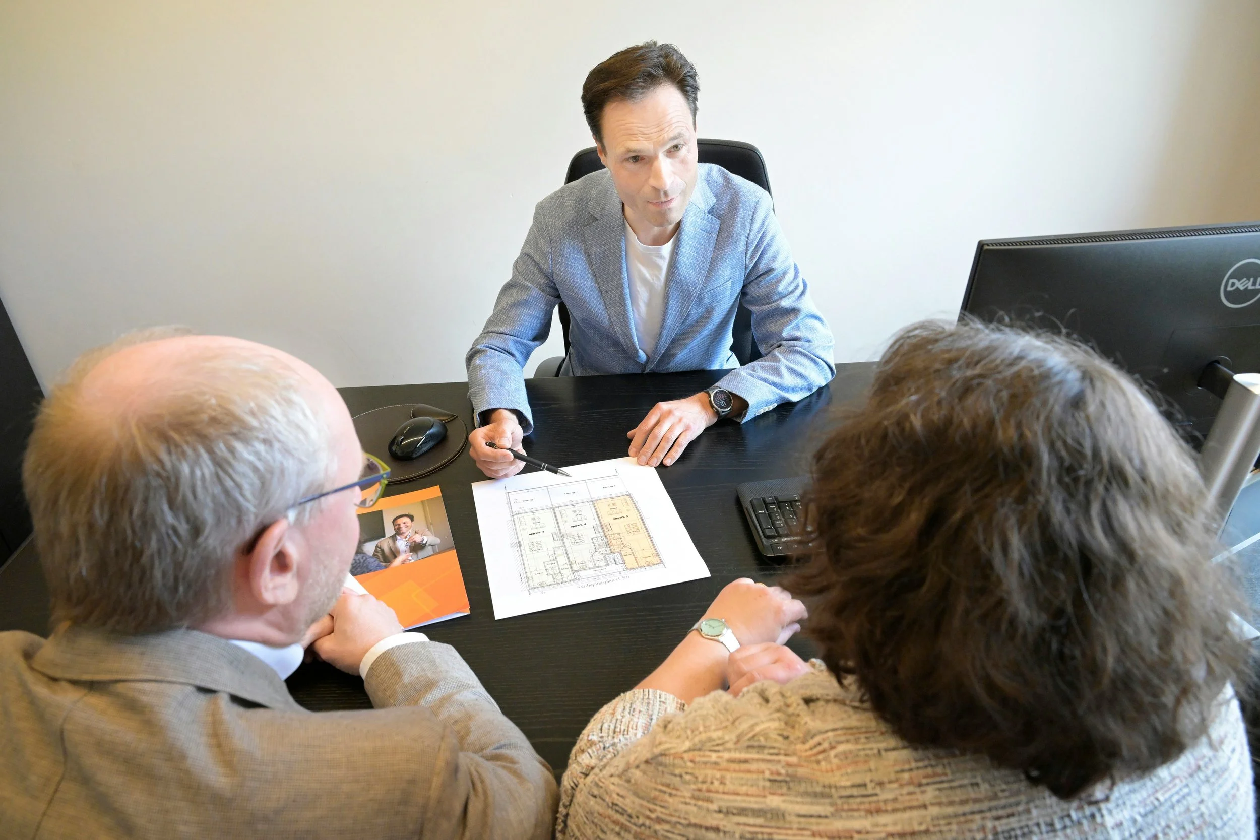 A man in a gray blazer and white shirt is speaking to a group of people in an office setting. The table has documents, a computer mouse, a book, and papers on it.