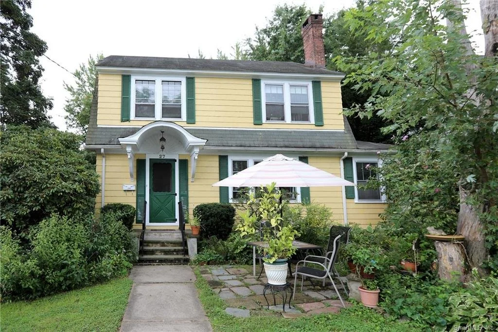 A yellow two-story house with green shutters and a dark green front door. There is a small set of stairs leading up to the front entrance, with bushes on either side. In the front yard, there is a patio with outdoor furniture, including a table, chairs, an umbrella, and potted plants, surrounded by trees and greenery.