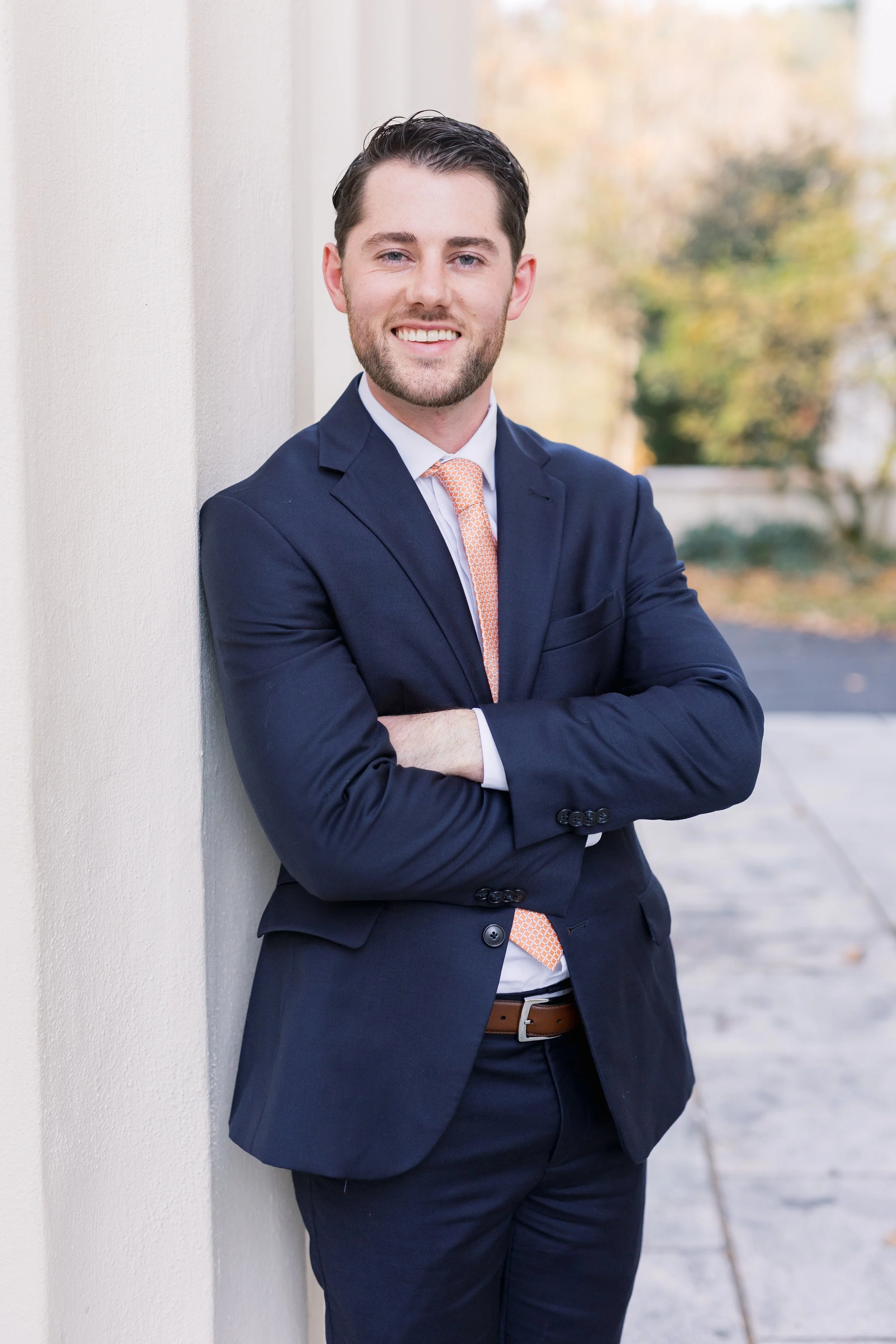 A young man with dark hair, light skin, and a beard, dressed in a navy blue suit, white shirt, and orange patterned tie, smiling and posing arm crossed while leaning against a white wall outdoors with blurred trees in the background.