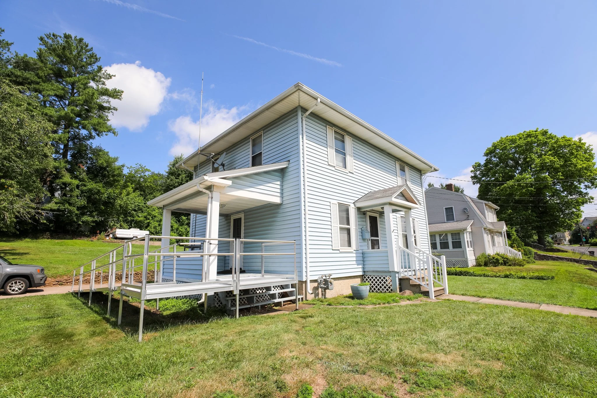 A light blue, two-story house with white shutters and a front porch with a ramp, situated in a grassy yard with trees and neighboring houses in the background.