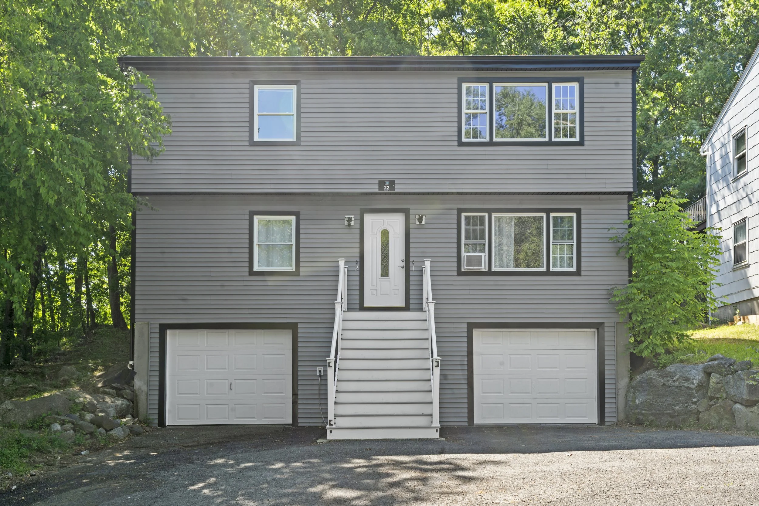 A modern, gray, two-story house with a front staircase, two garage doors on the ground level, and several windows, surrounded by green trees.
