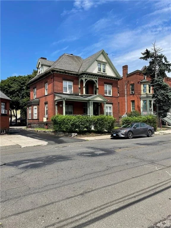 A large, historic three-story brick house with a green gabled roof, front porch, and decorative trim, situated on a city street with a grey car parked nearby, under a blue sky with some clouds.