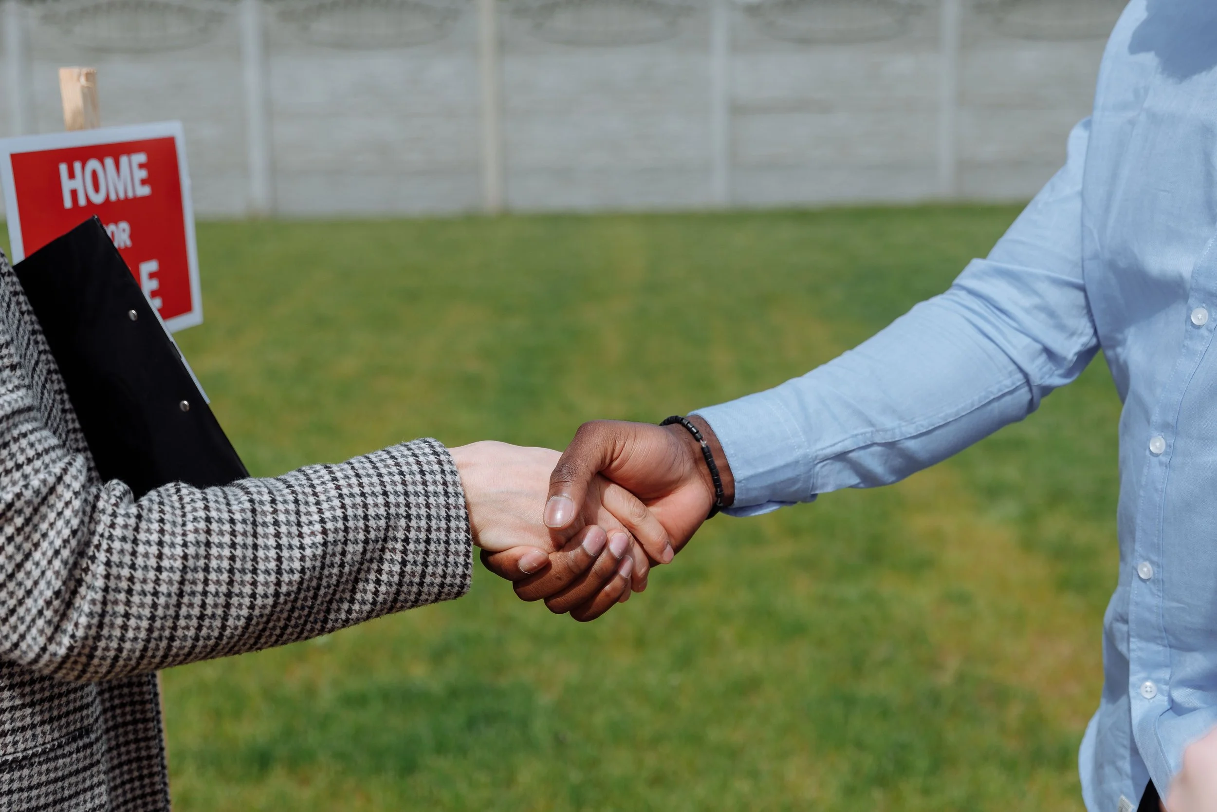 Two people shaking hands outdoors with a green lawn and a sign that reads 'Home' in the background.