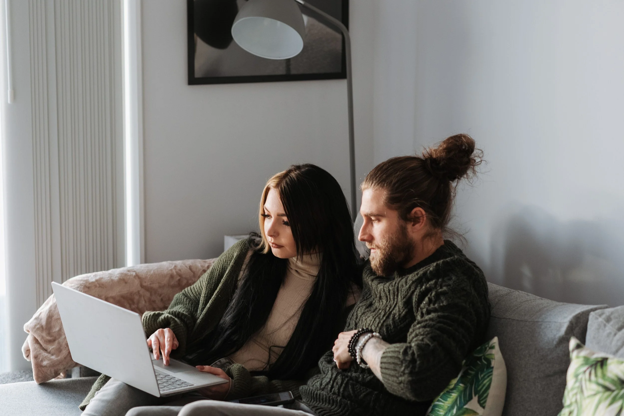 A man and a woman sitting close together on a beige couch, looking at a laptop screen. The woman has long dark hair and is wearing a beige turtleneck with a dark green sweater. The man has long hair tied in a bun and a beard, wearing a dark sweater and bracelets. They appear focused, possibly working or watching something on the laptop. The room has white walls, a framed mirror, and a lamp in the background.