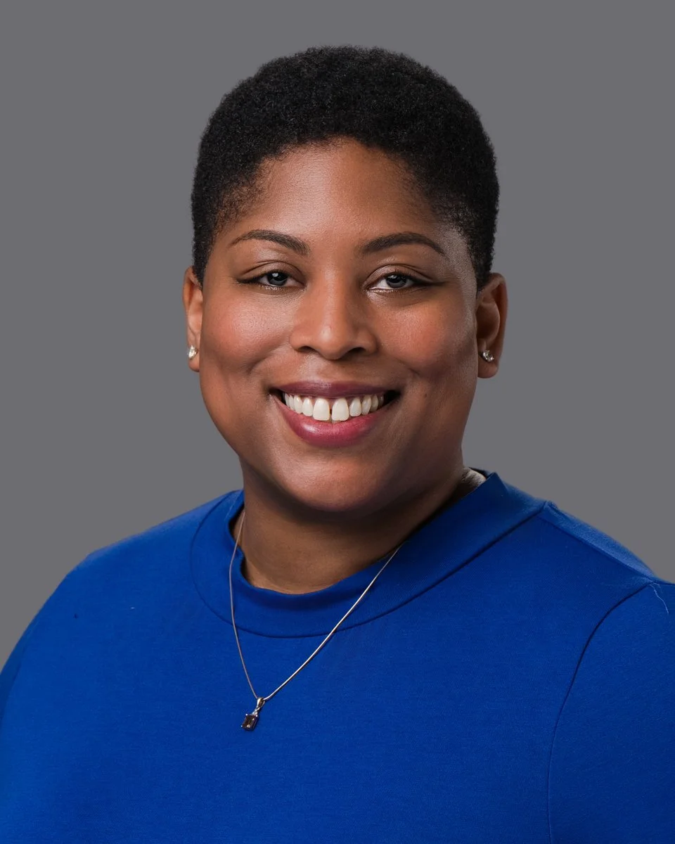 A portrait of a smiling African American woman with short black hair, wearing a blue top, earrings, and a necklace, against a gray background.