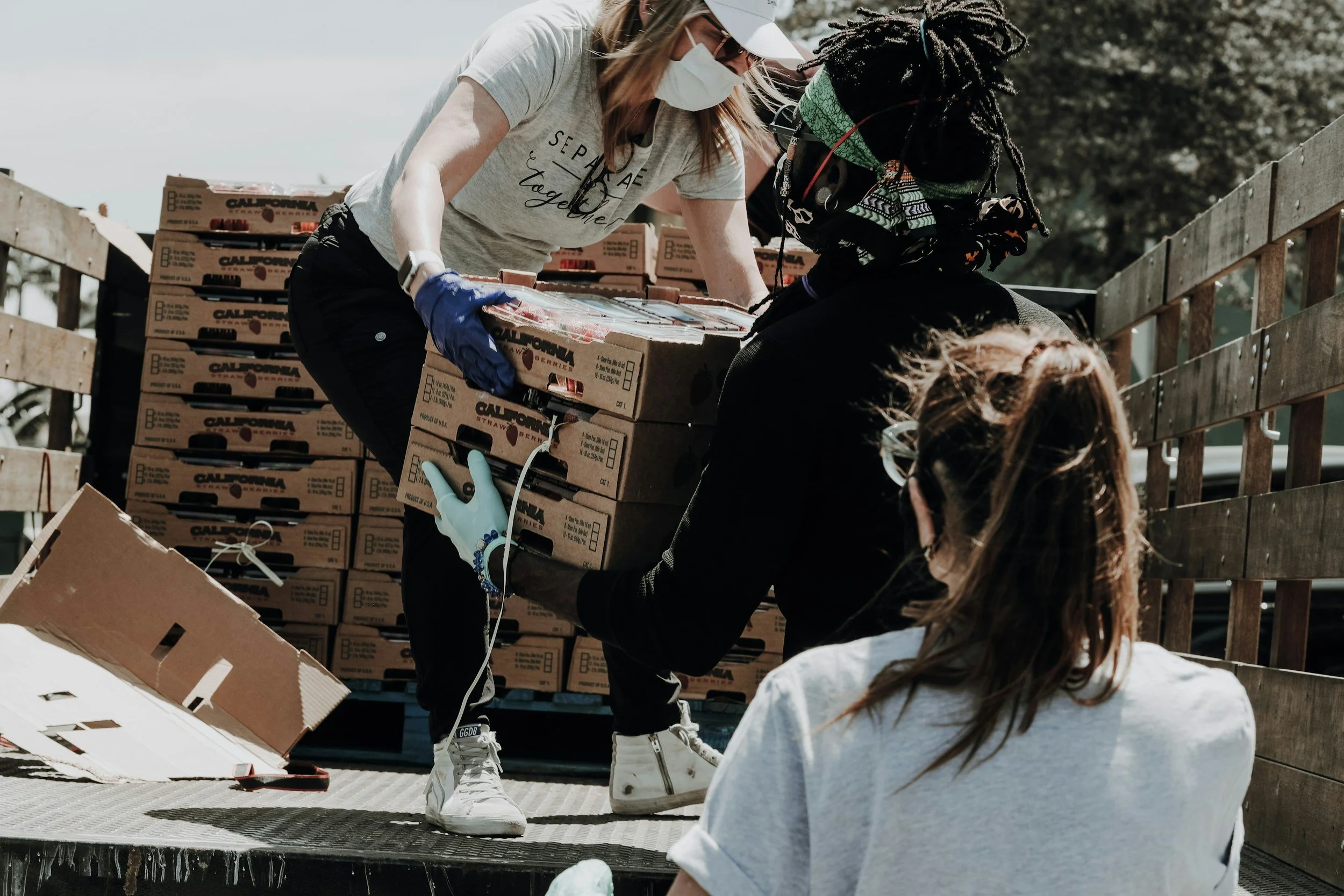 People working together unloading boxes labeled 'California Strawberries' from a truck at an outdoor location.