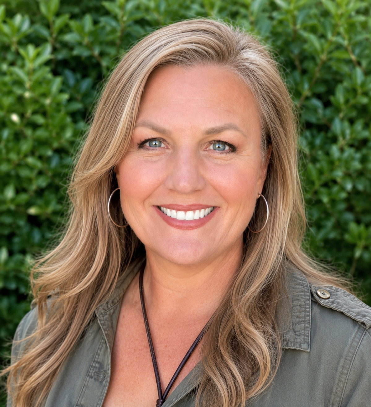 A smiling woman with long blonde hair, wearing hoop earrings, a gray jacket, and a necklace, standing outdoors in front of green foliage.