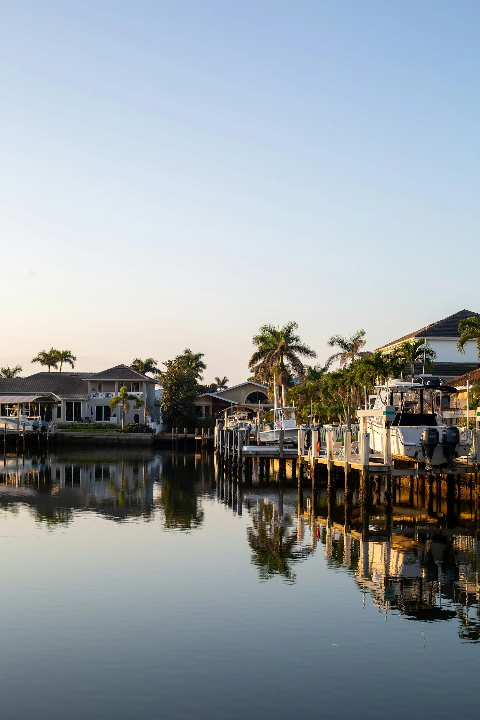 A peaceful waterfront scene with boats docked along a marina, houses with palm trees, and a calm water reflection during sunset or early evening.