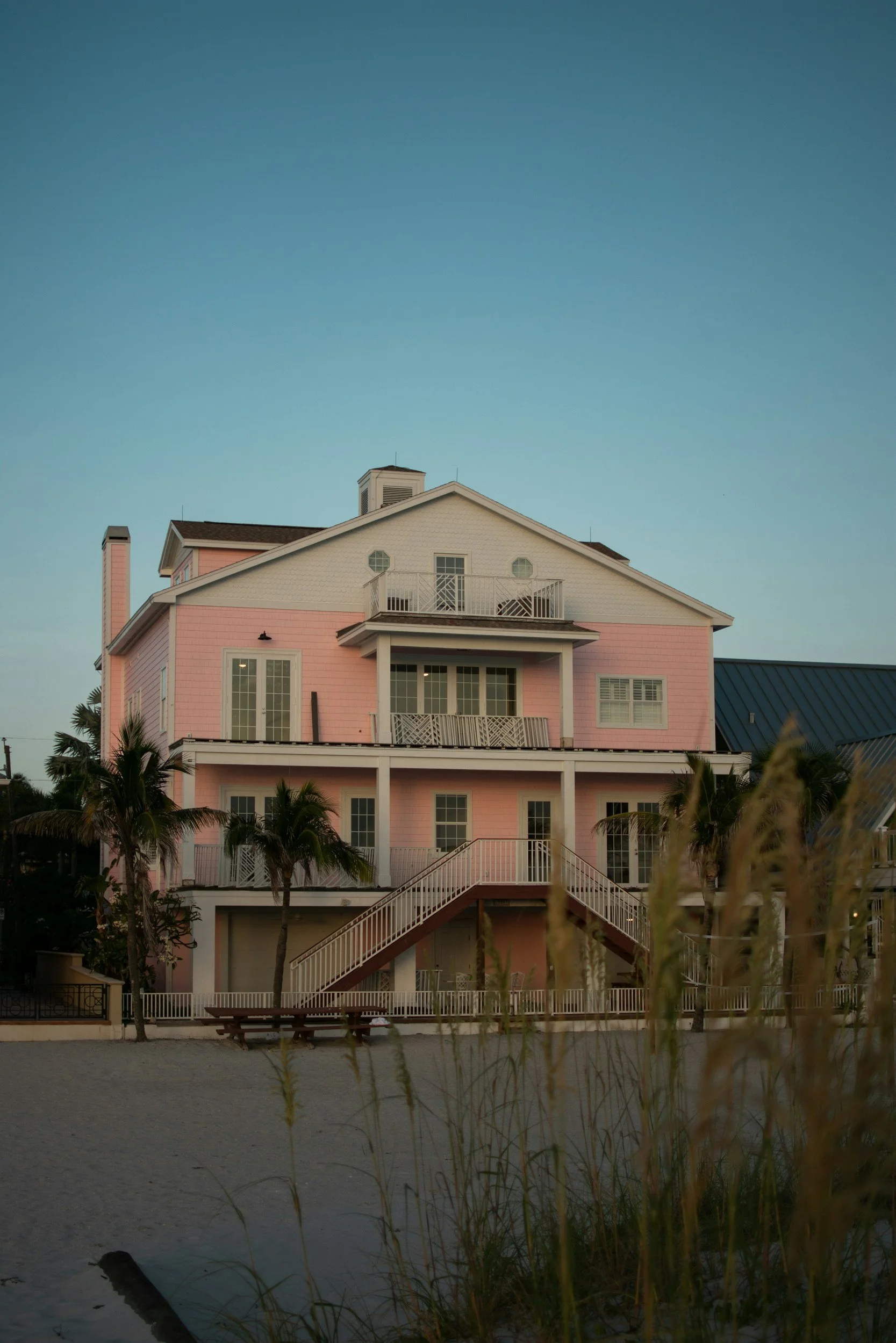 Pink house with multiple stories, balconies, and stairs leading up to the entrance, surrounded by palm trees and a body of water in the foreground, under a clear blue sky at sunset.