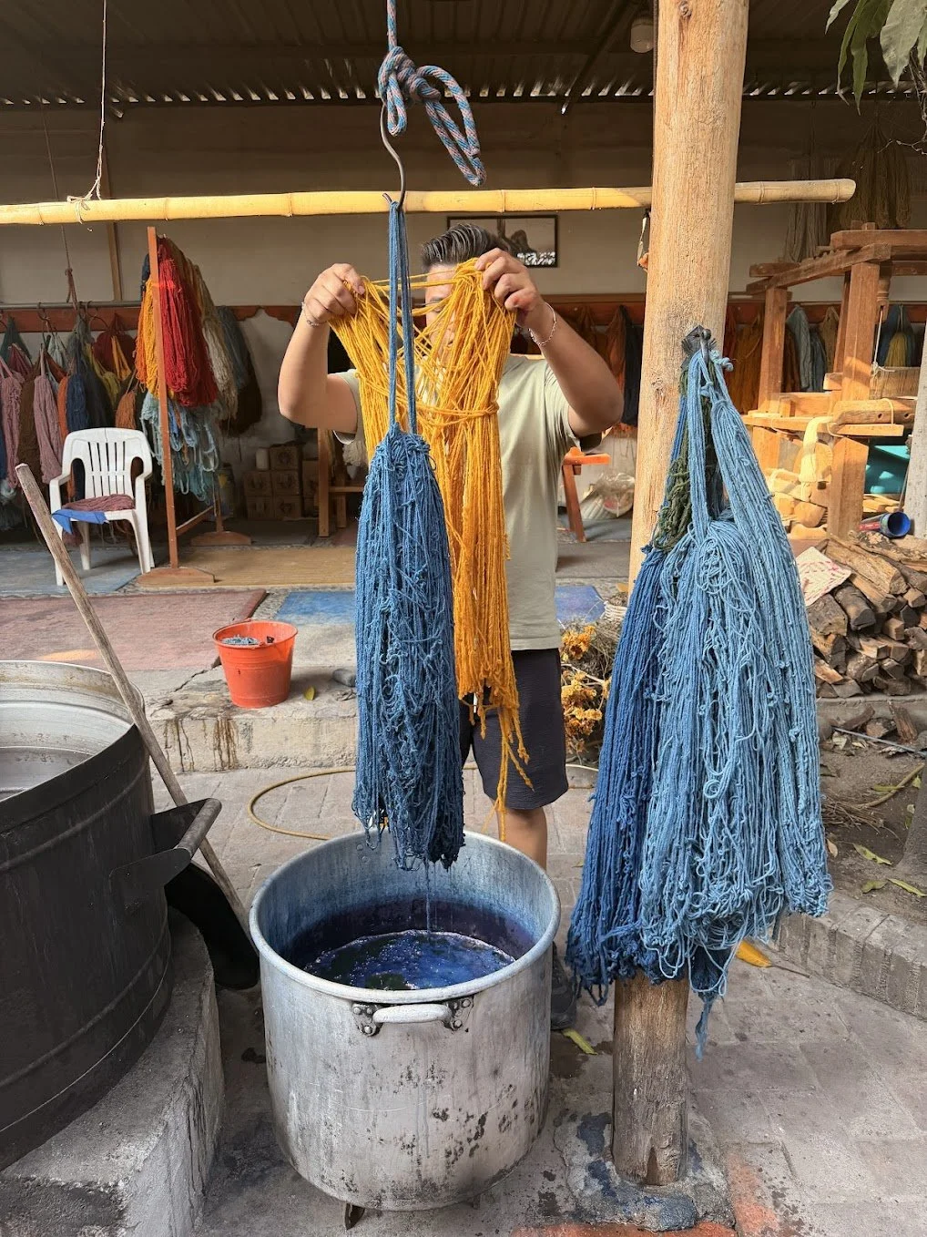 Person hanging yellow and blue yarn to dry over a metal tub filled with natural dye, in an outdoor workshop with shelves and colorful yarns in the background.