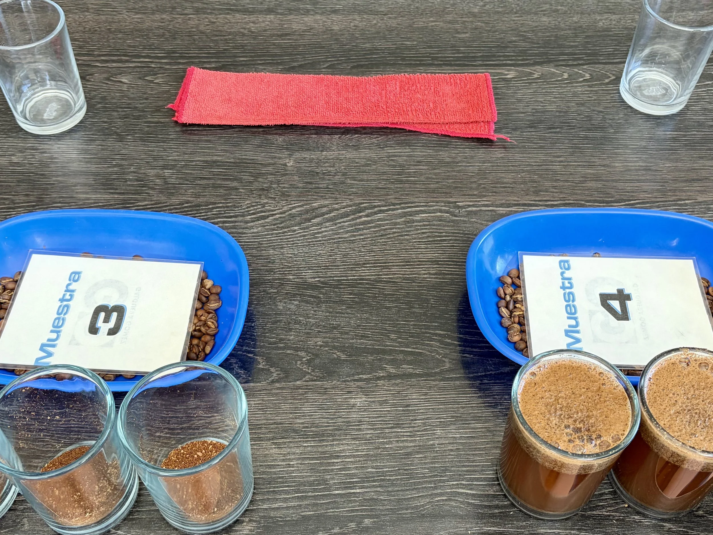 Several glasses filled with a frothy, brown beverage on a dark wooden table, alongside blue bowls containing coffee beans and cards with numbers and text.