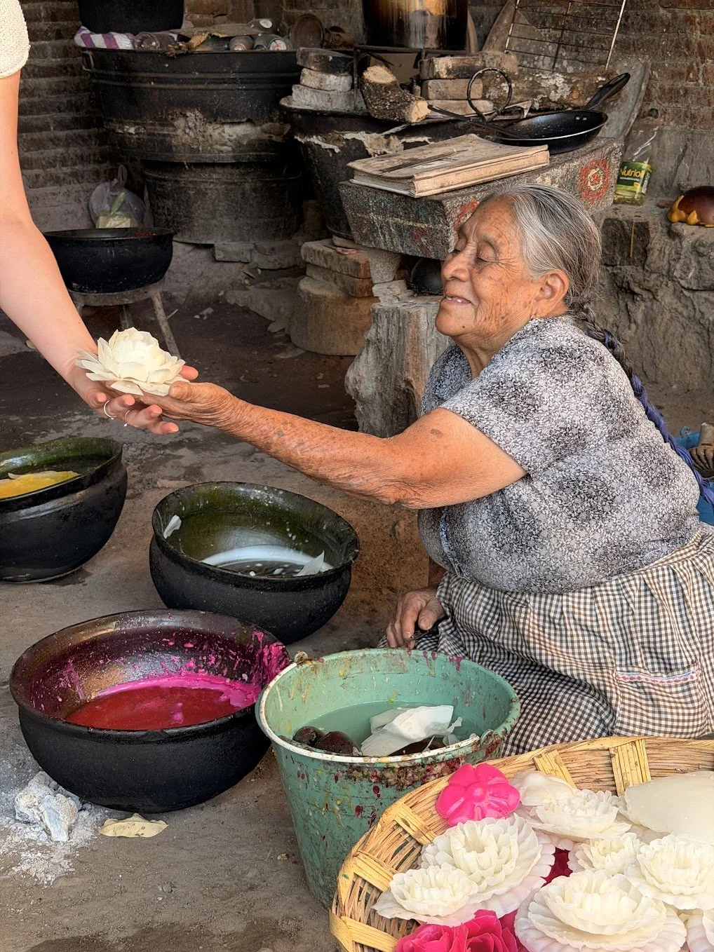 An elderly woman in a checkered skirt and gray blouse smiling as she receives a white water lily flower from a person outside the frame, in a rustic outdoor setting with bowls and buckets of colored dye and handmade paper flowers.