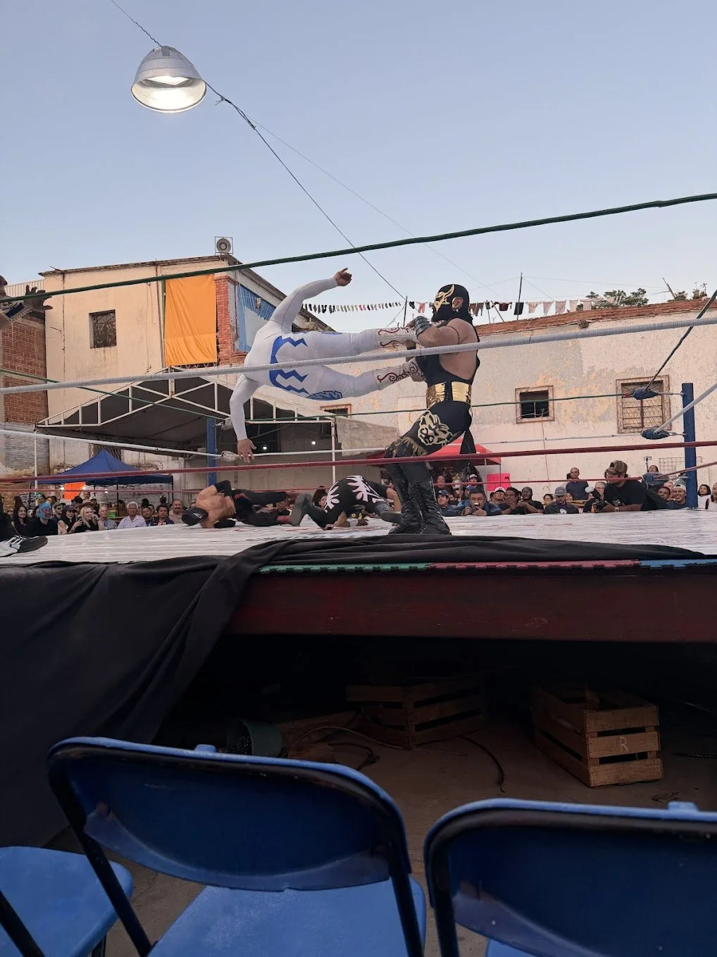A lucha libre wrestling match taking place outdoors with a large crowd watching. Two masked wrestlers are engaged in a move; one in a white and blue costume flying towards the other in a black and gold costume. The ring is surrounded by ropes and sit
