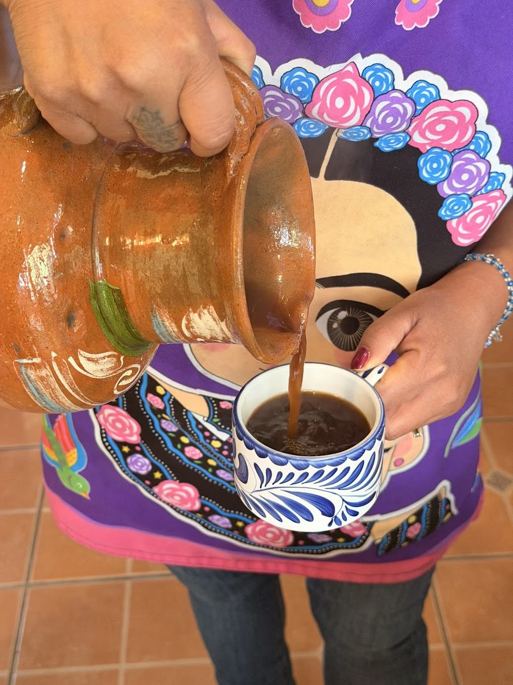 Person pouring coffee from a clay pitcher into a patterned mug, wearing a colorful dress with a floral and Frida Kahlo design.