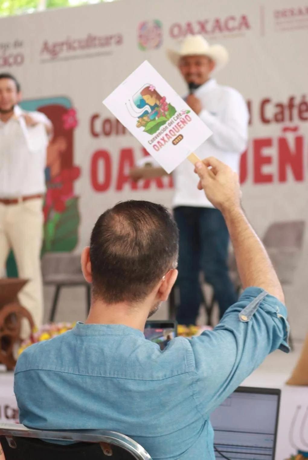 A man is holding a sign with a colorful cartoon drawing and the words "CONVENCIÓN DEL CAFÉ OAXAQUEÑO" in front of a stage where two men are standing and speaking. The background features a banner with the word "OAXACA" and related decorations.