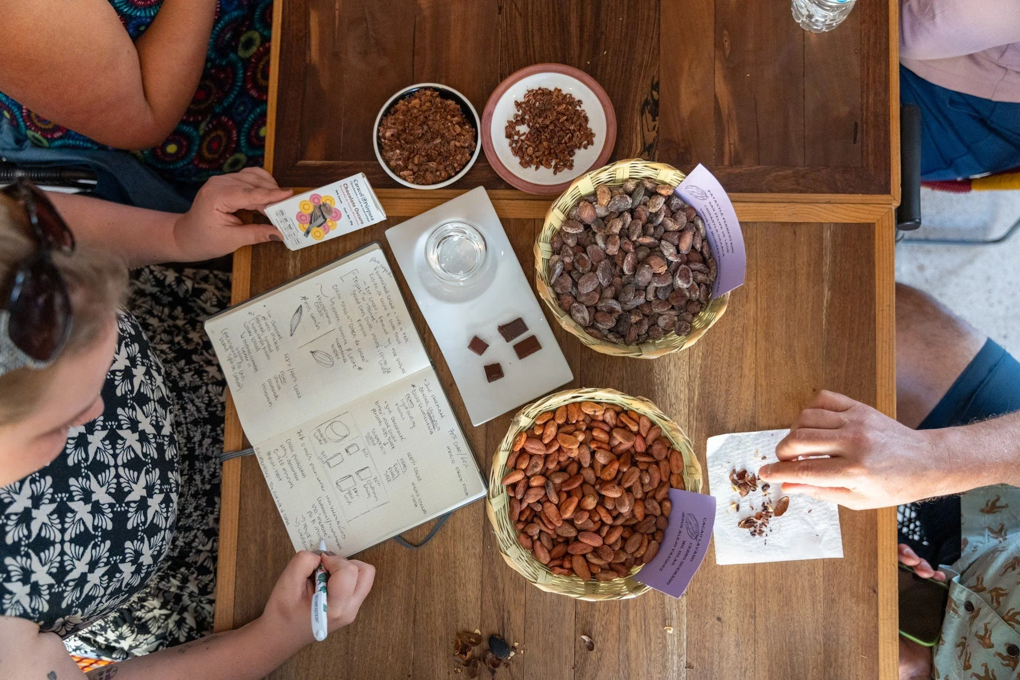 Overhead view of a wooden table with baskets of cocoa beans, bowls of crushed cocoa nibs, a glass of water, a tasting sheet, and people sampling chocolate.