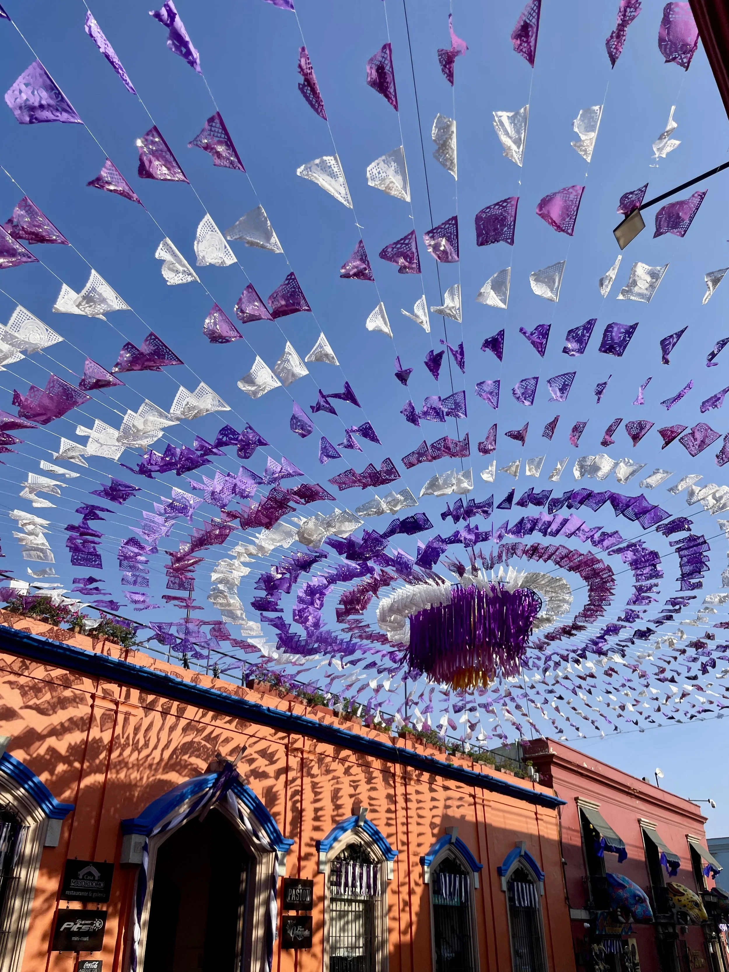 Colorful decorative banners in purple and white hang in the sky above a pink building with arched windows and a red roof on a sunny day.