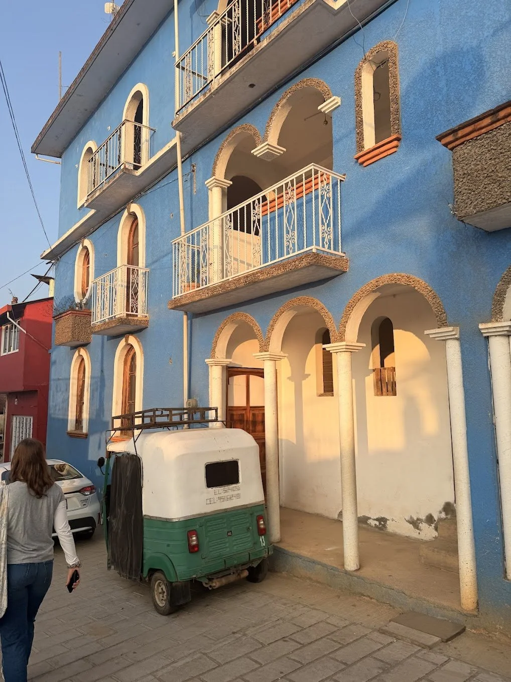 Colorful blue building with arched and rectangular windows, small balconies with decorative railings, and white pillars at the entrance. A woman walks below the building, and a small green and white vehicle is parked in front.
