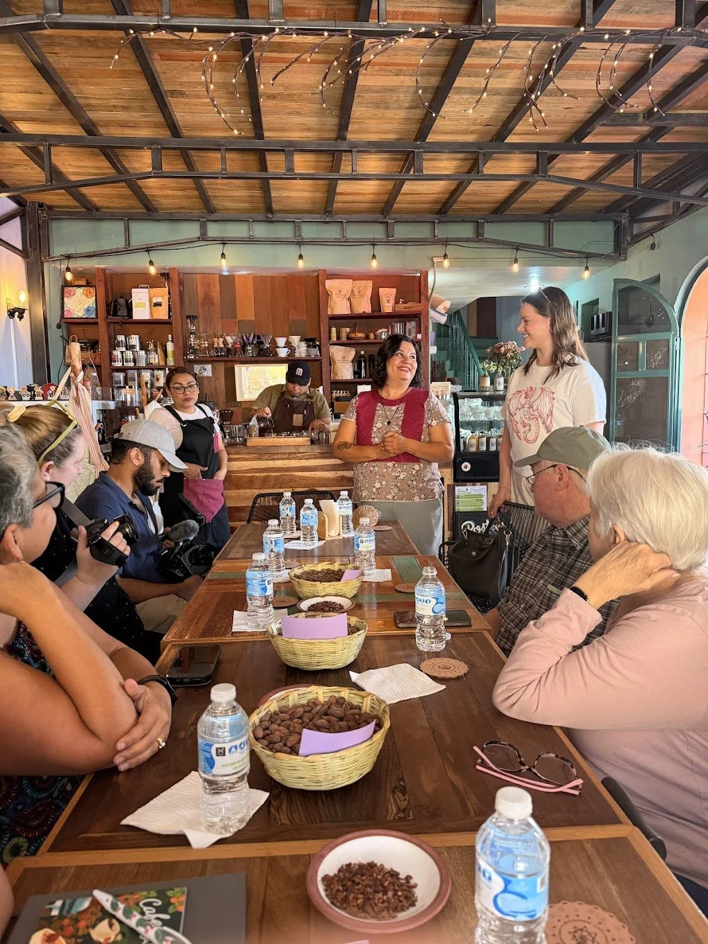A group of people sitting around a wooden table with water bottles and baskets of samples, listening to two women and a man speaking inside a rustic cafe or tasting room.