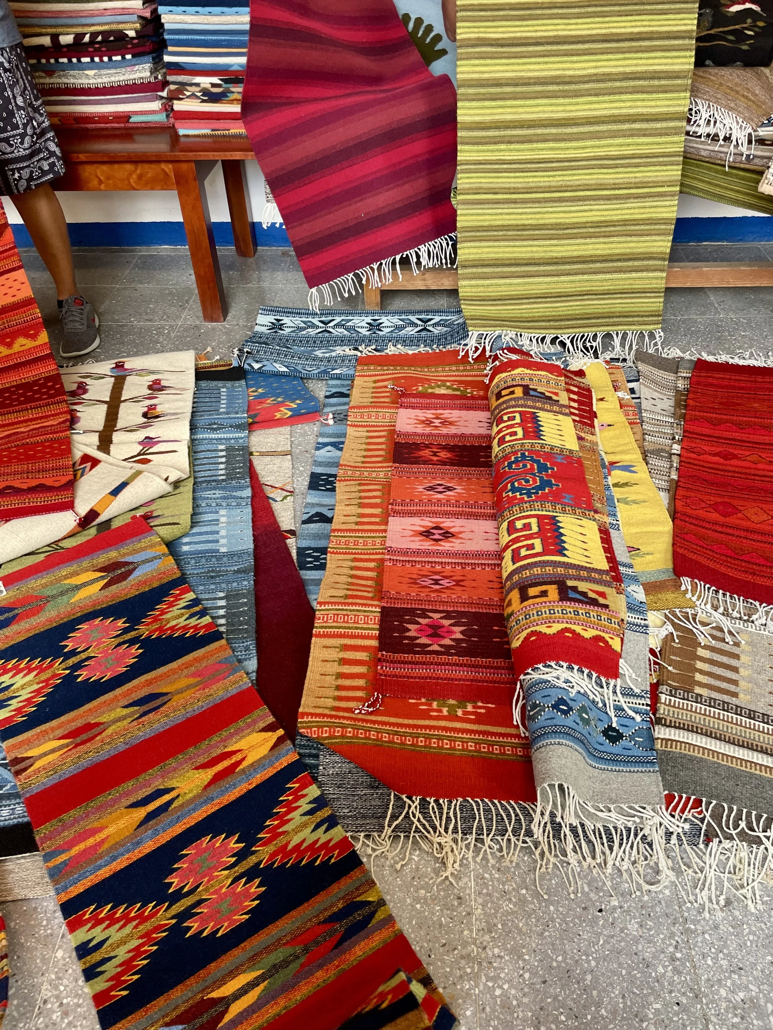 Display of colorful woven rugs with various geometric and traditional patterns, some rolled and others laid flat on the floor, with a background of shelves filled with stacks of fabric.