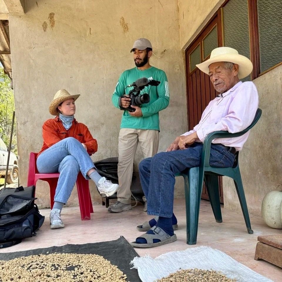 A young woman in a tan hat, orange shirt, and jeans sitting on a pink chair, a man in a green shirt holding a video camera. An older man in a white hat and light pink shirt sitting on a green plastic chair nearby. The setting appears to be outdoors with a rustic wall behind them, and there are some bags on the ground and some coffee beans laid out on cloths in front of them.