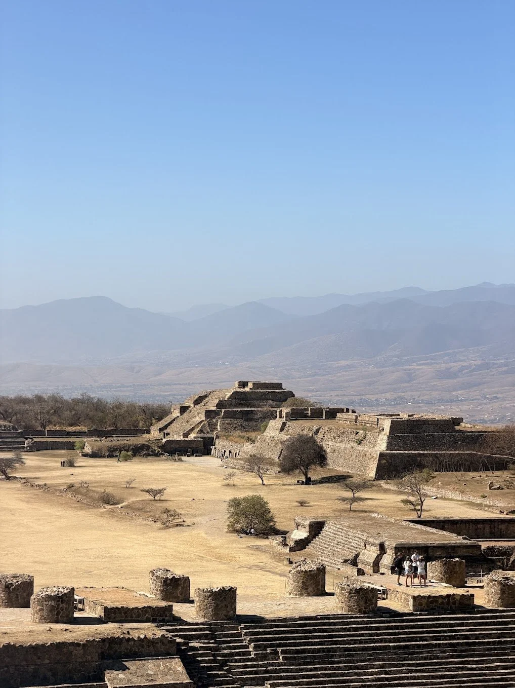 Ancient stepped pyramids and stone structures in a dry landscape with sparse trees, set against a backdrop of distant mountains under a clear blue sky.