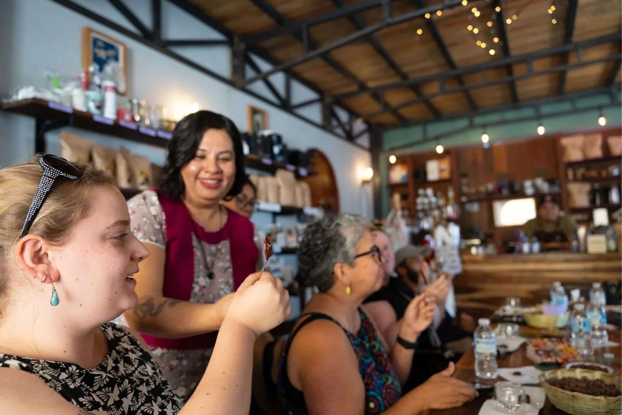 People gathered around a communal table at a café, enjoying chocolate samples, rustic setting with wooden shelves, string lights, and a high ceiling.