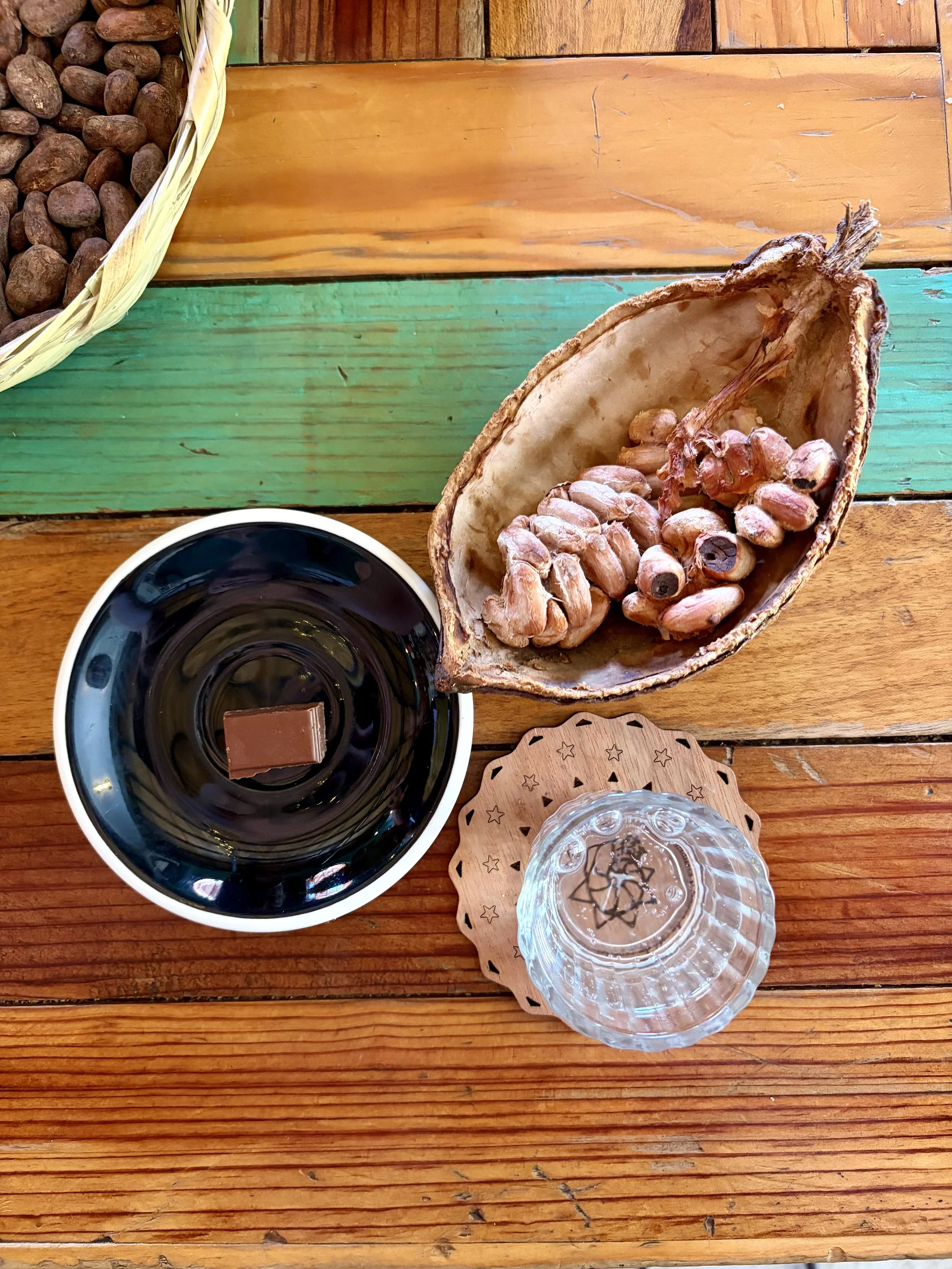 A wooden table with a basket of roasted cacao seeds, a saucer with a piece of chocolate on top, a cacao pod opened in half, and a glass of water with a coaster.