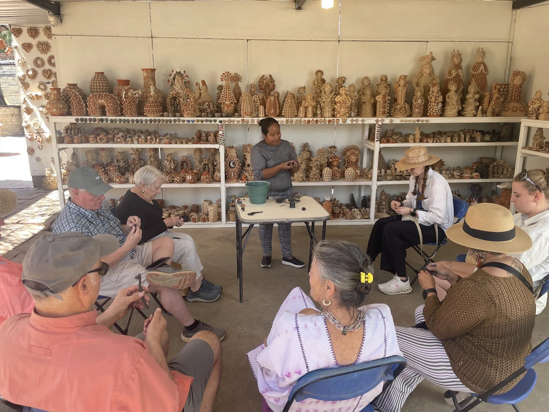 A group of people sitting on chairs in an indoor space, watching a woman presenting and demonstrating how to make decorative pottery figures, with shelves full of similar pottery art in the background.
