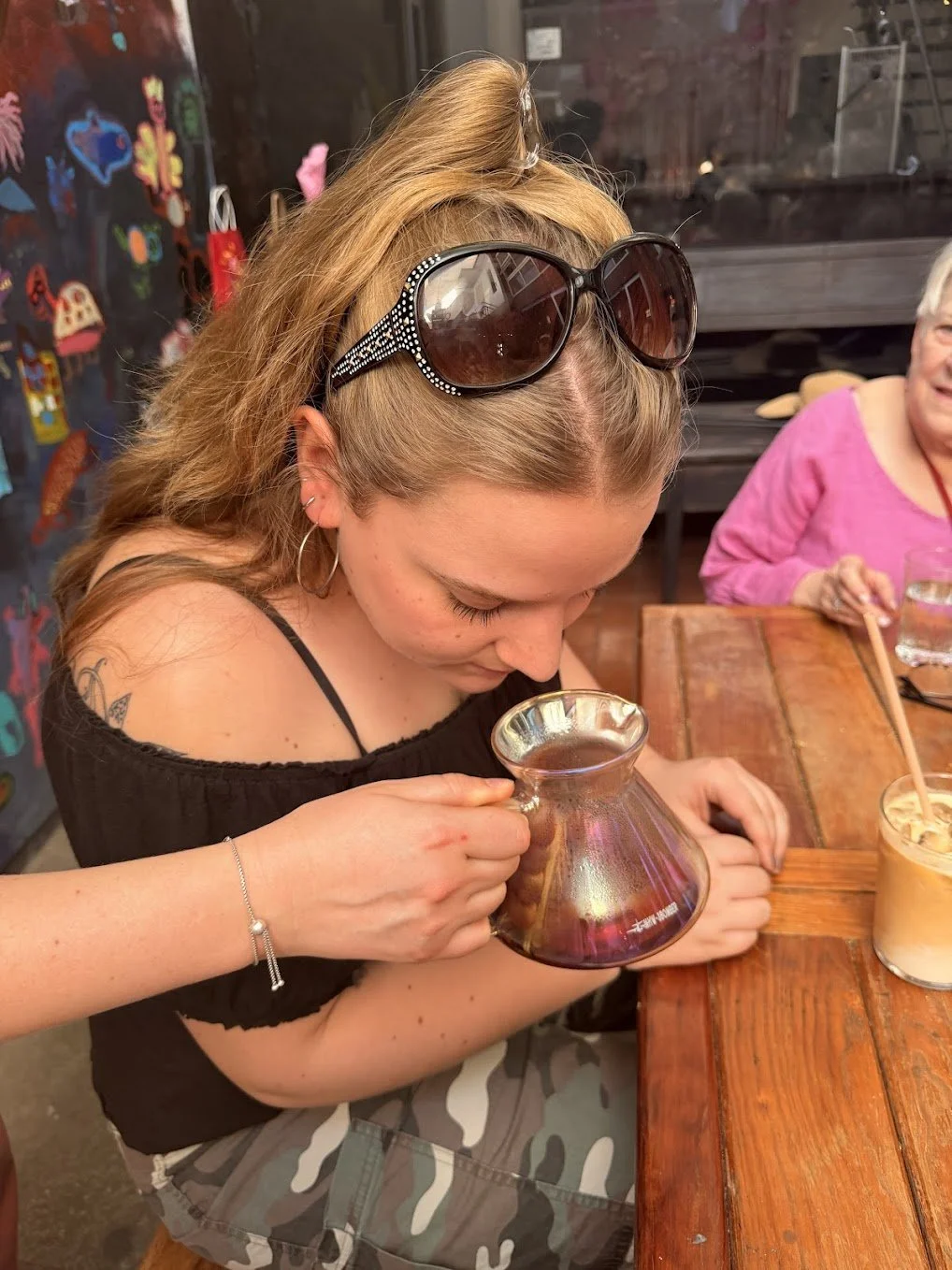 A young woman with glasses on her head holding a small, iridescent glass pitcher while sitting at a wooden table in a restaurant. She has a tattoo on her shoulder and is wearing a black off-shoulder top. An older woman in a pink shirt is visible in t